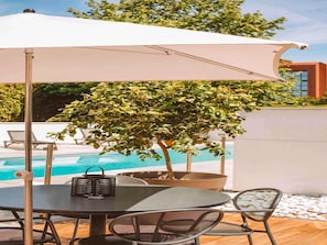 A poolside area with a table and chairs under a white umbrella, surrounded by a tree and a modern building in the background.