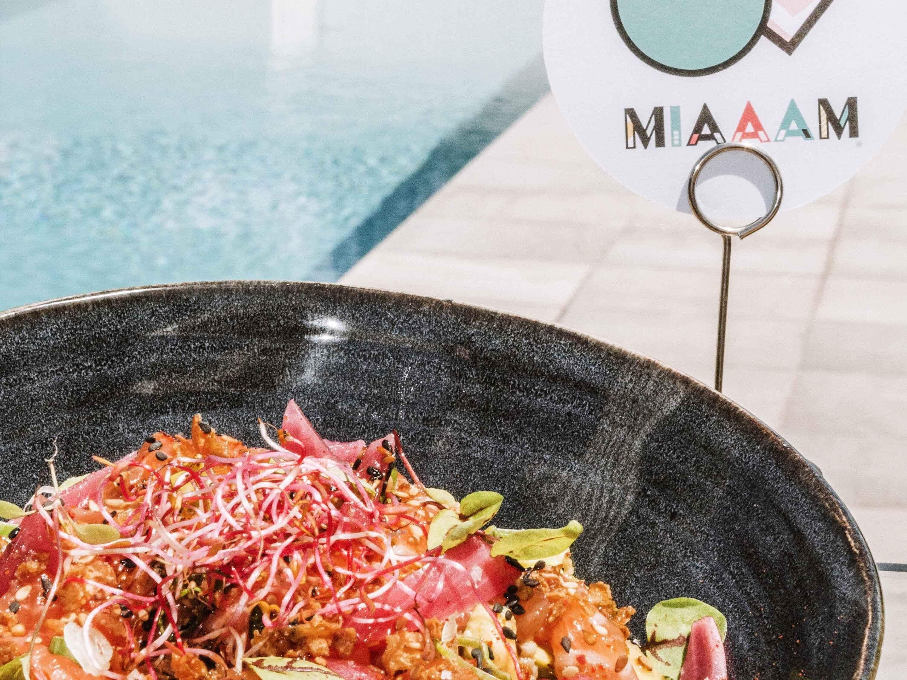 A black bowl of salad with various toppings, a pool in the background, and a sign with 'MAAM' visible.