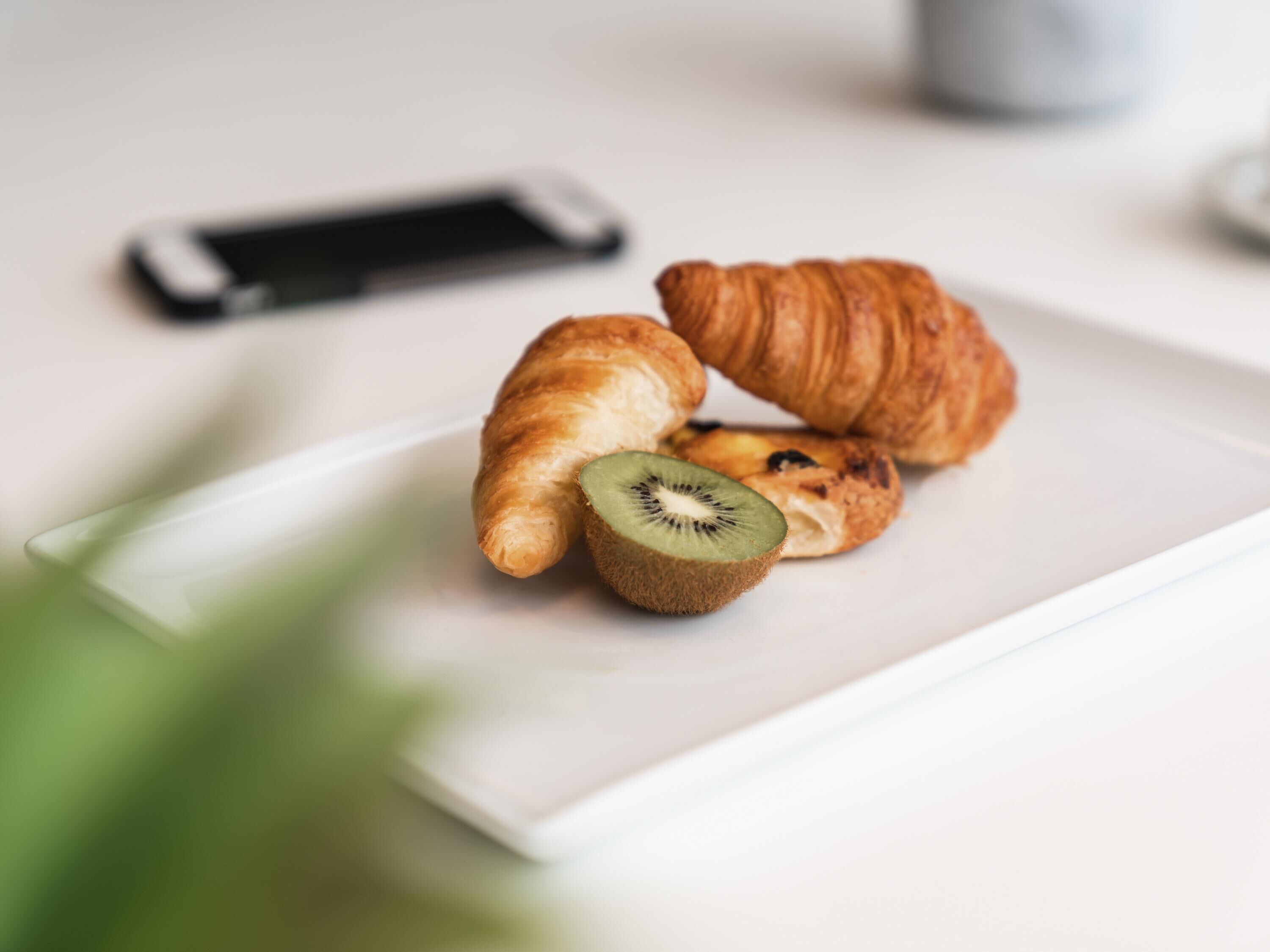 A plate with croissants and a kiwi slice on a white table with a smartphone in the background.