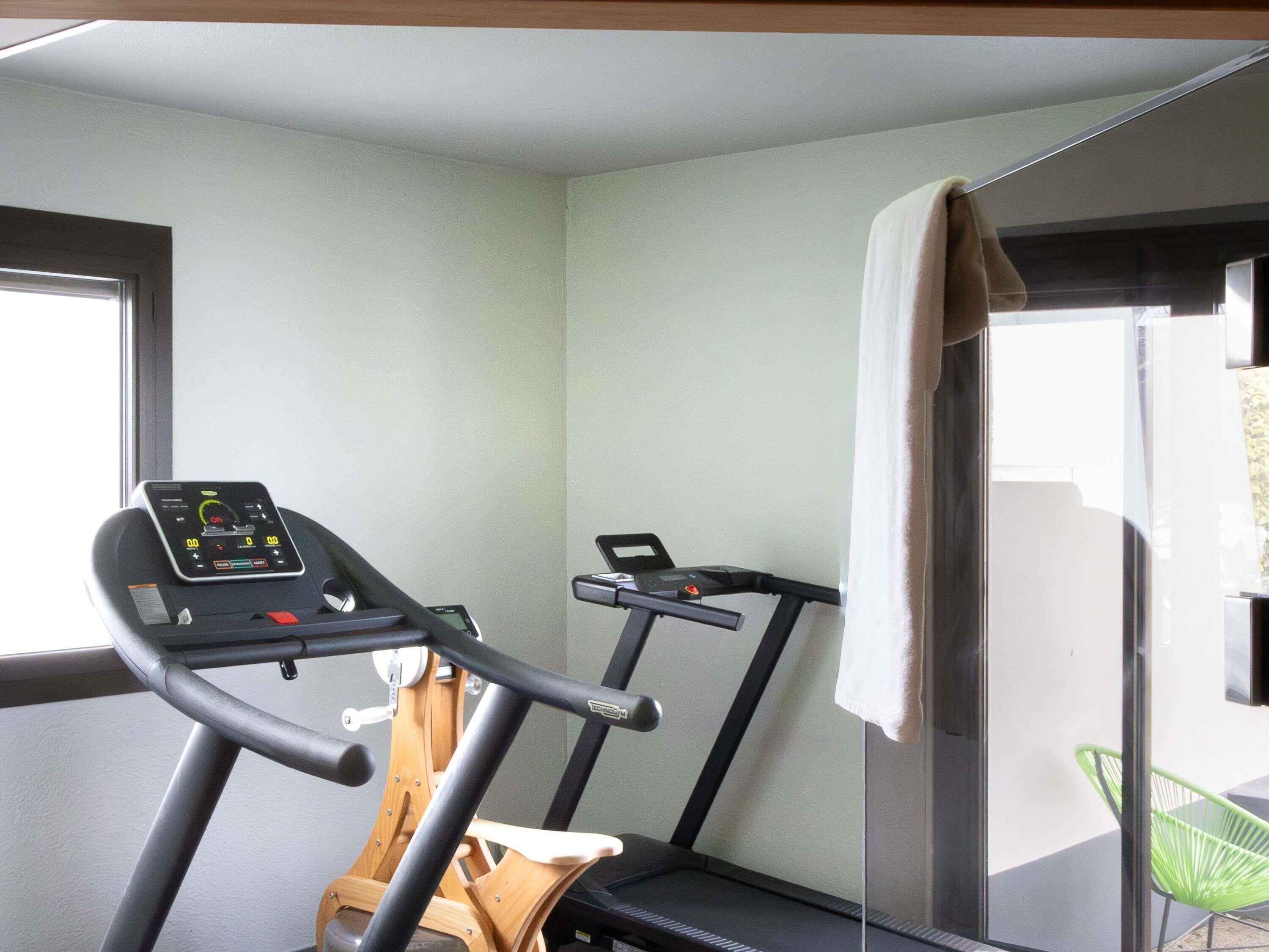 Una habitación con una cinta de correr y una bicicleta estática, una ventana con vistas, y una silla verde.