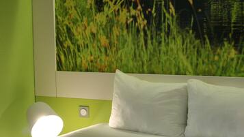 A bed with white bedding and pillows, a wall-mounted lamp, and a large framed picture of a field.