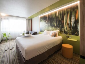 Habitación de hotel con cama, una silla verde, una mesita pequeña y un mural grande con temática forestal.