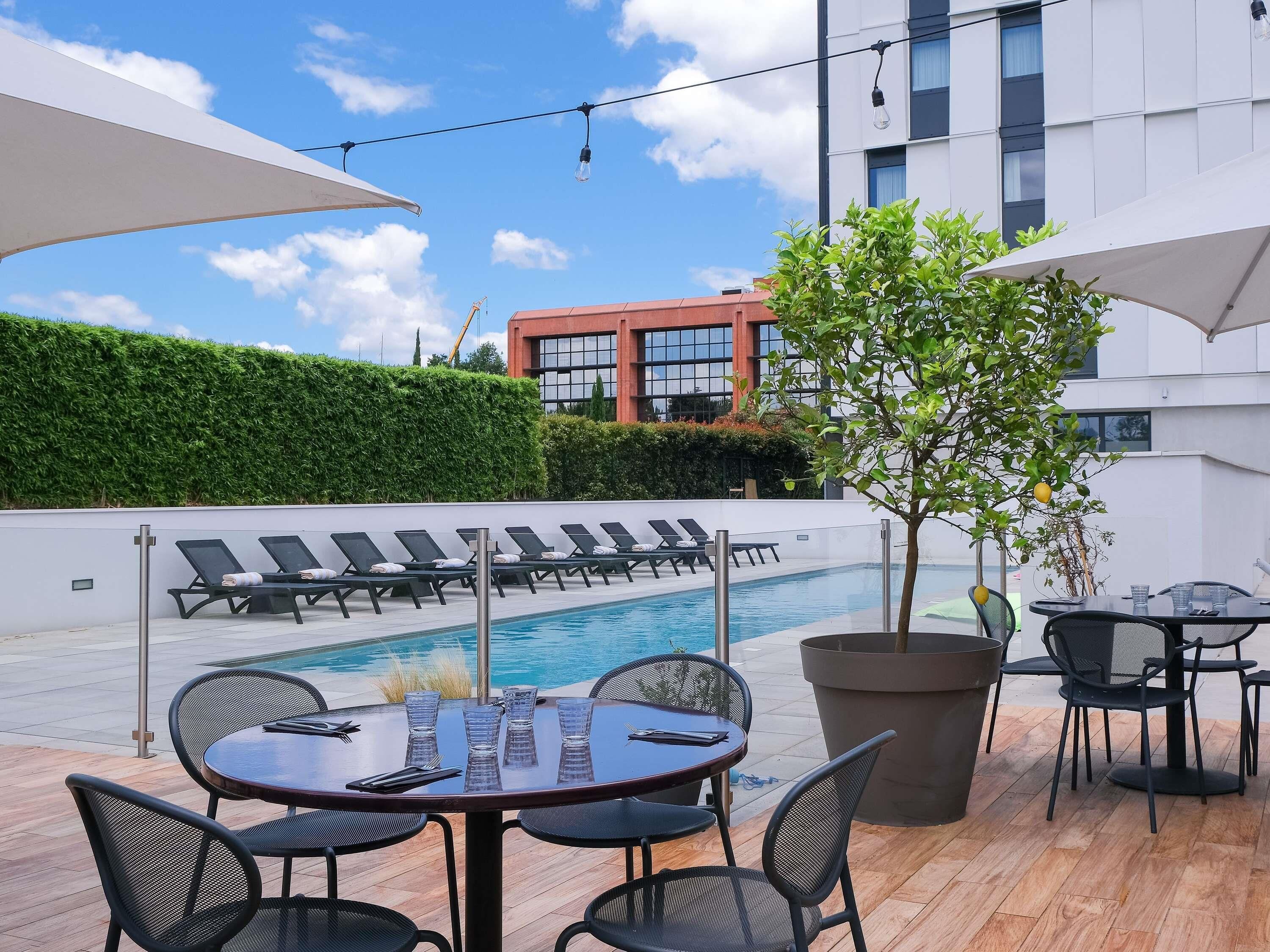 A poolside area with lounge chairs, a table with chairs, umbrellas, and a building in the background.