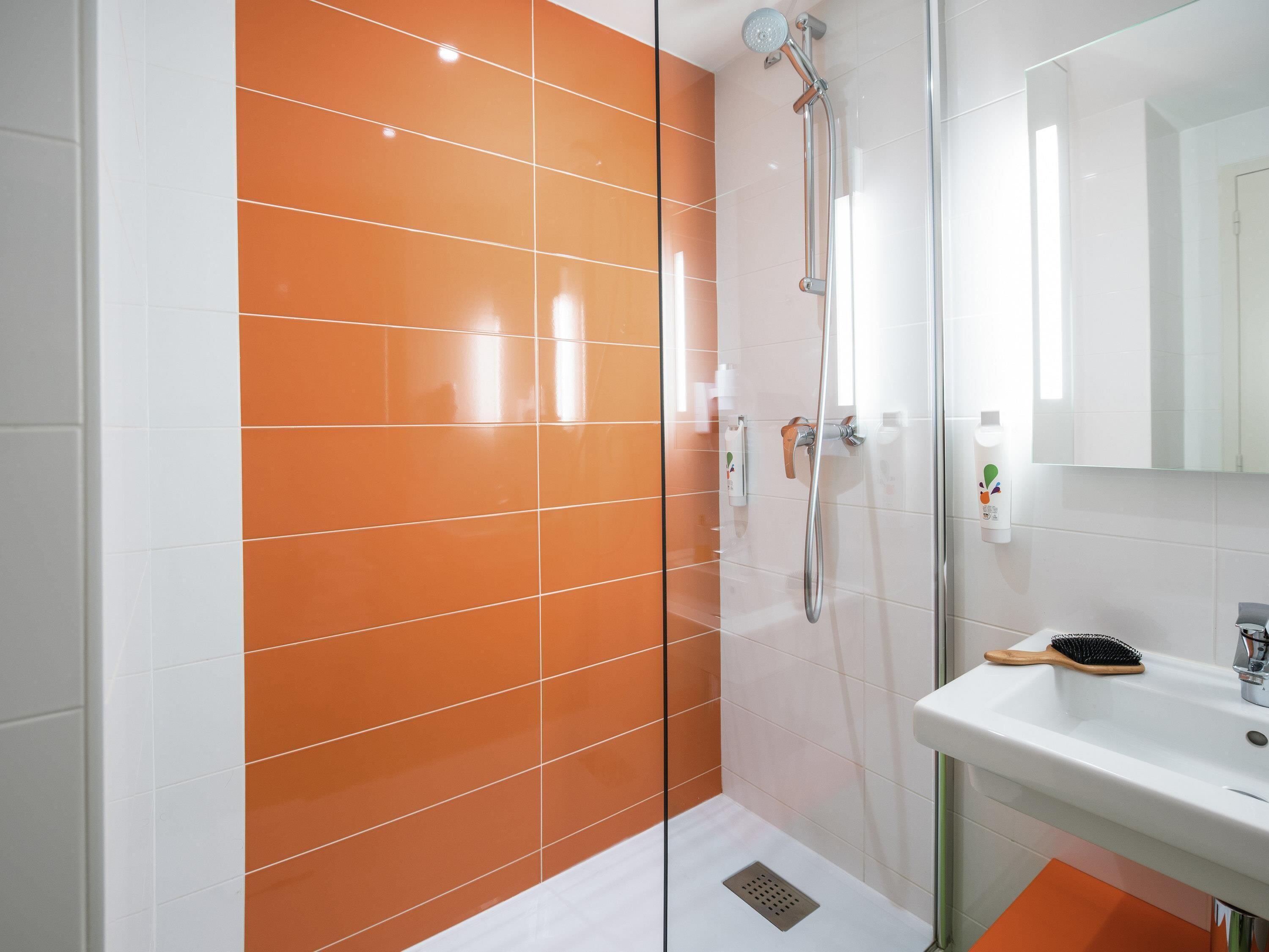 A modern bathroom with orange and white tiles, a glass shower enclosure, a sink with a mirror, and a toothbrush holder.