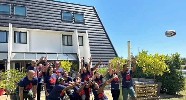 A group of people outdoors in front of a building with a sloped roof and multiple windows.