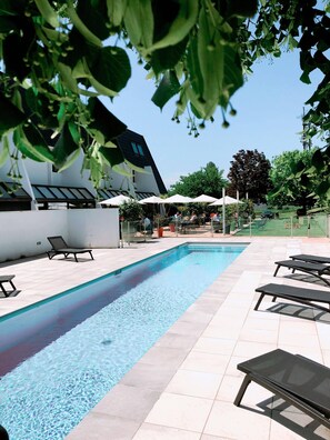 Piscina con sillones de descanso y sombrillas, rodeada de un edificio blanco y vegetación.