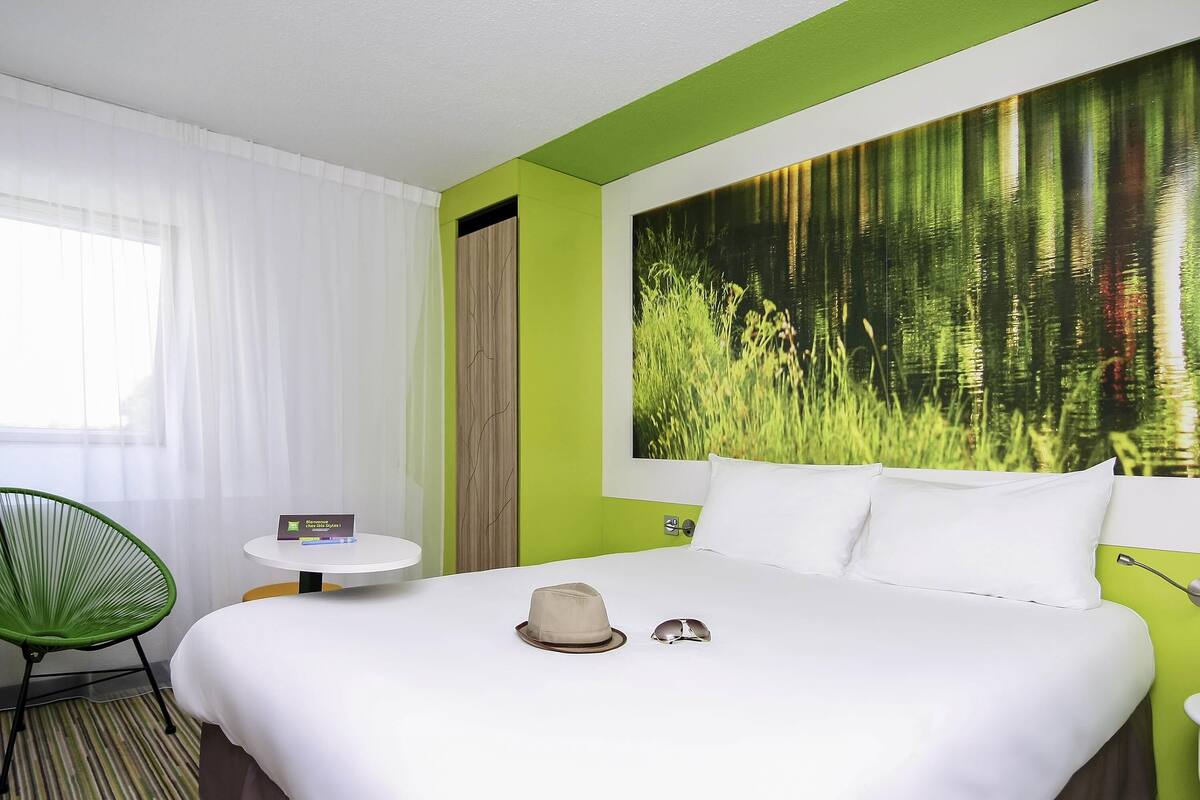 A hotel room with a bed, a green chair, a small table with a book, and a large nature-themed wall mural.