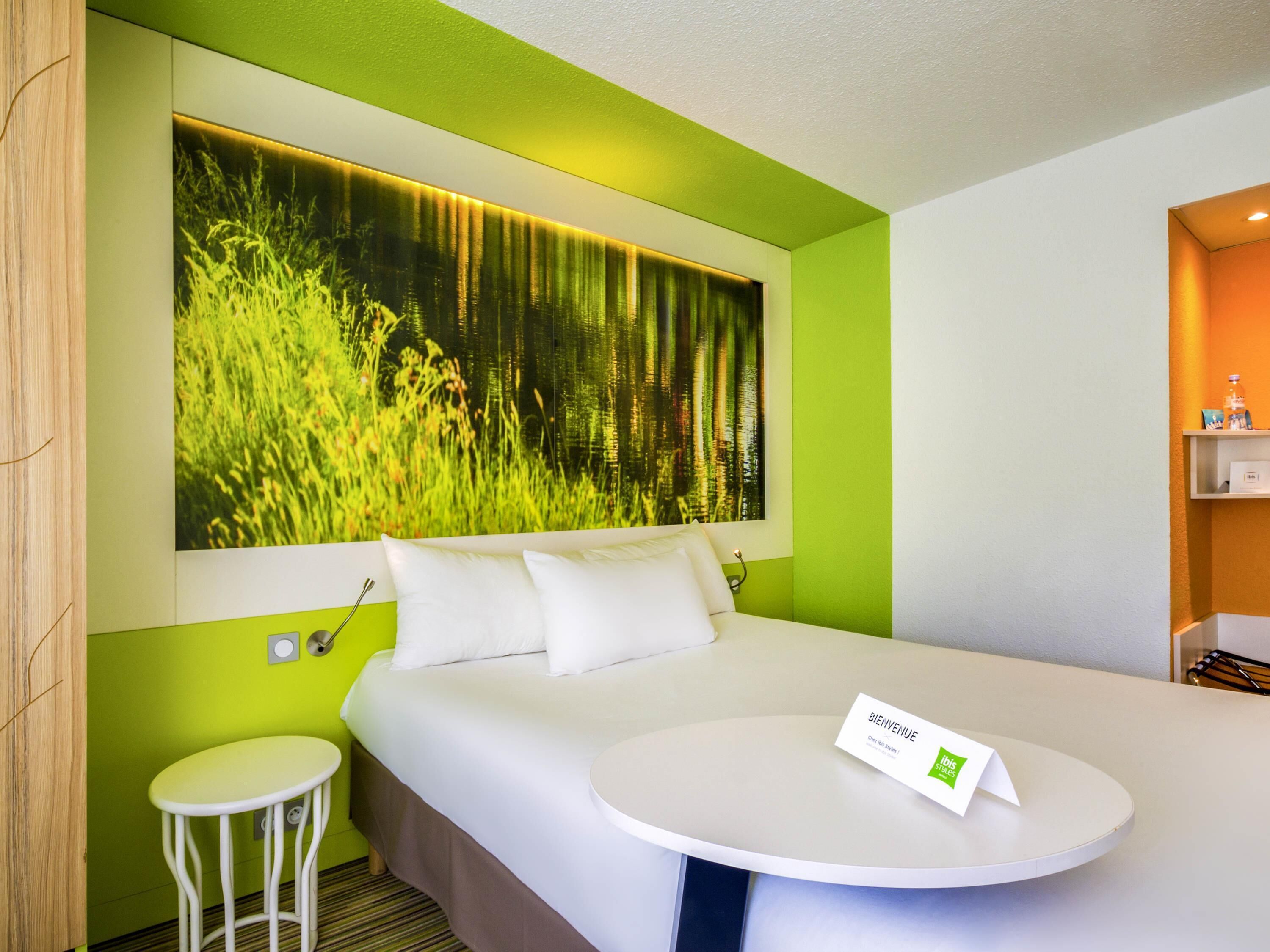 Una habitación de hotel moderna con una cama grande, una pequeña mesa redonda, una pared de acento verde y una obra de arte de temática natural sobre la cama.