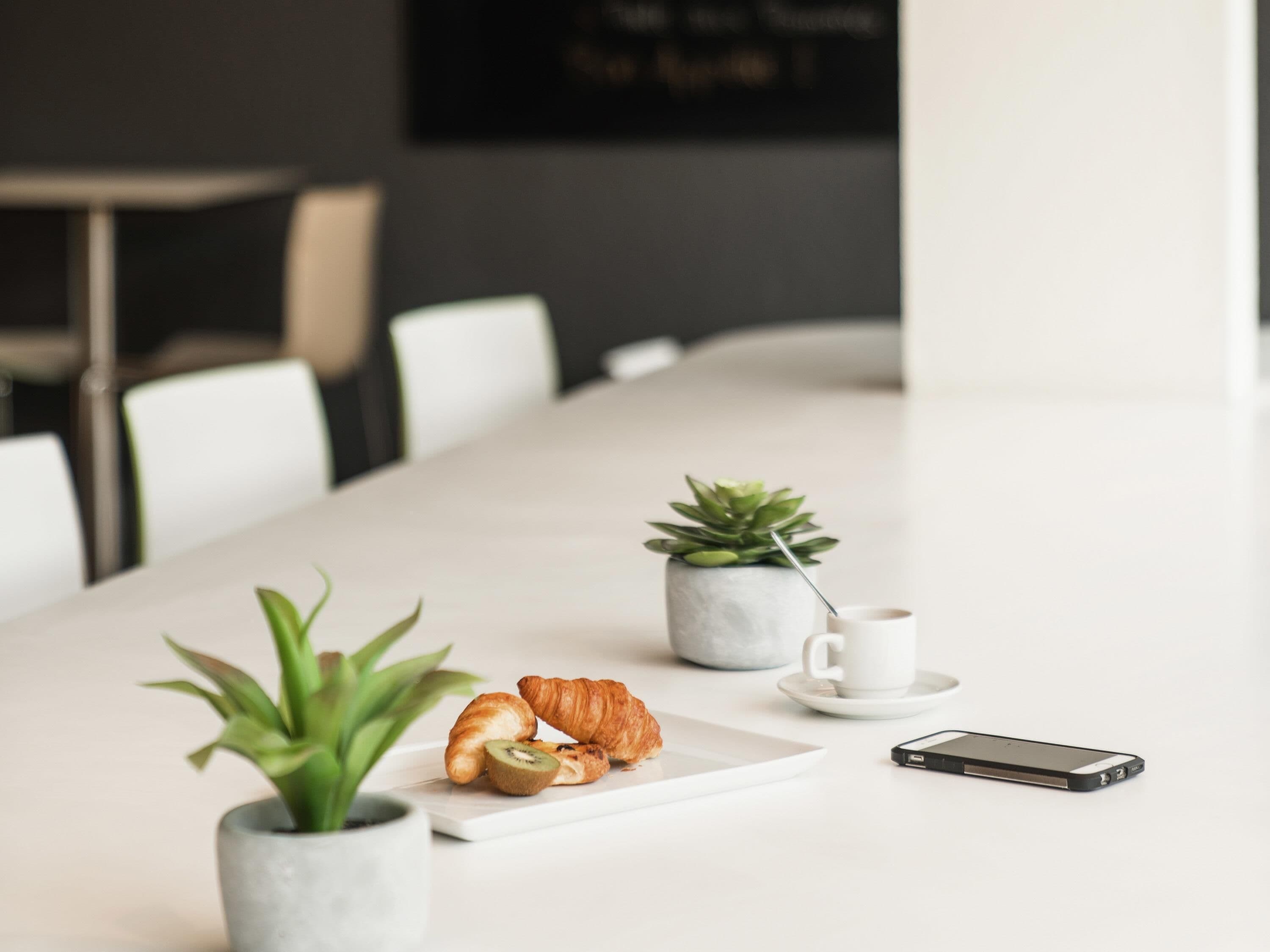 Una larga mesa con un croissant, una rebanada de kiwi, un teléfono inteligente y dos plantas en macetas.
