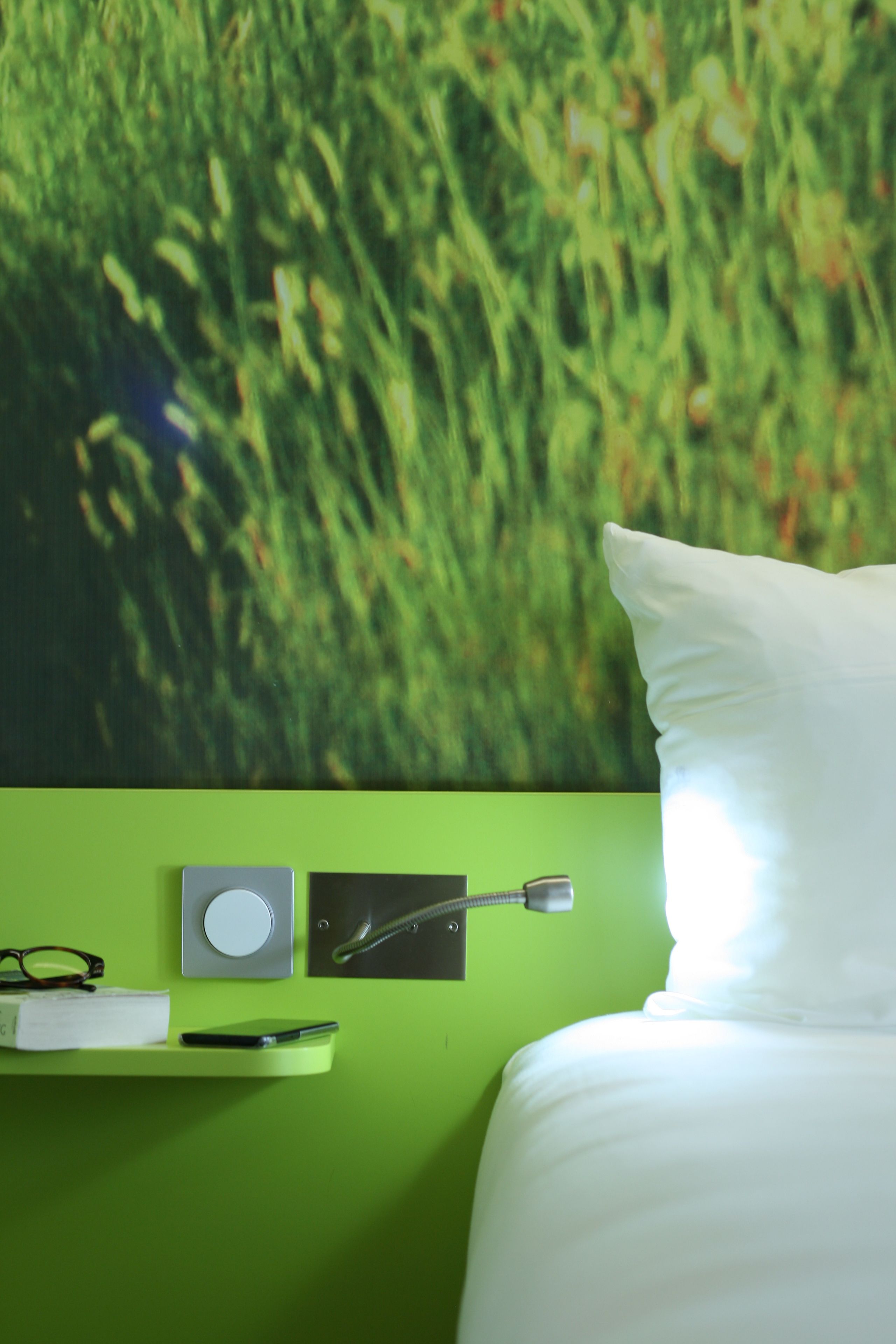 A room with a green wall, a white pillow, a bed, a shelf with a book, glasses, a smartphone, and a wall-mounted device.