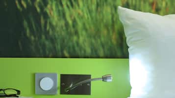 A room with a green wall, a white pillow, a bed, a shelf with a book, glasses, a smartphone, and a wall-mounted device.