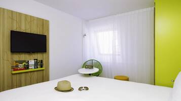 A hotel room with a bed, a television mounted on the wall, a small table with a chair, and a window with curtains.