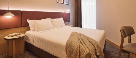 A hotel room with a large bed, a bedside table with a lamp, a chair, and a window with curtains.