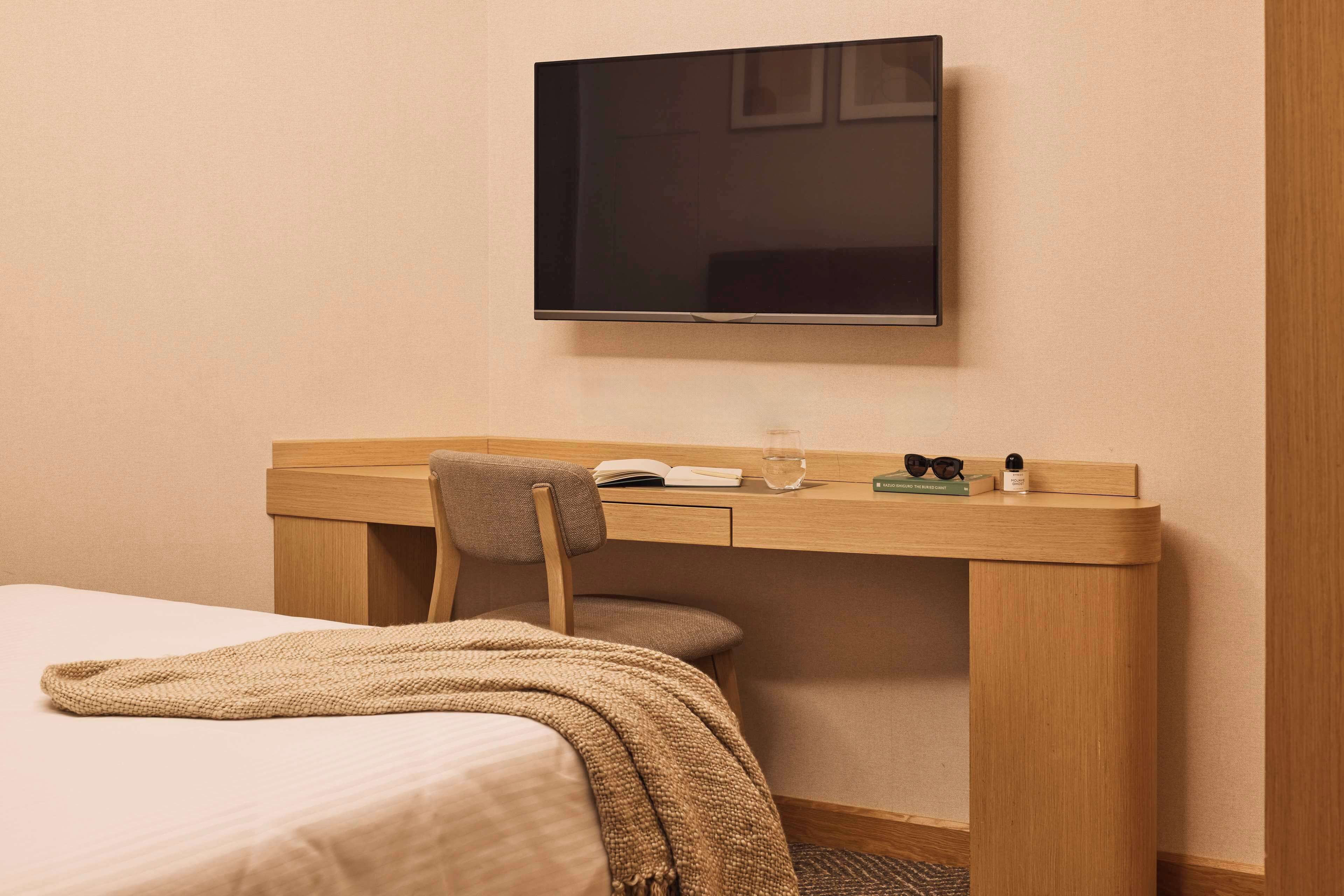 A hotel room with a bed, a desk with a chair, a television mounted on the wall, and a window.