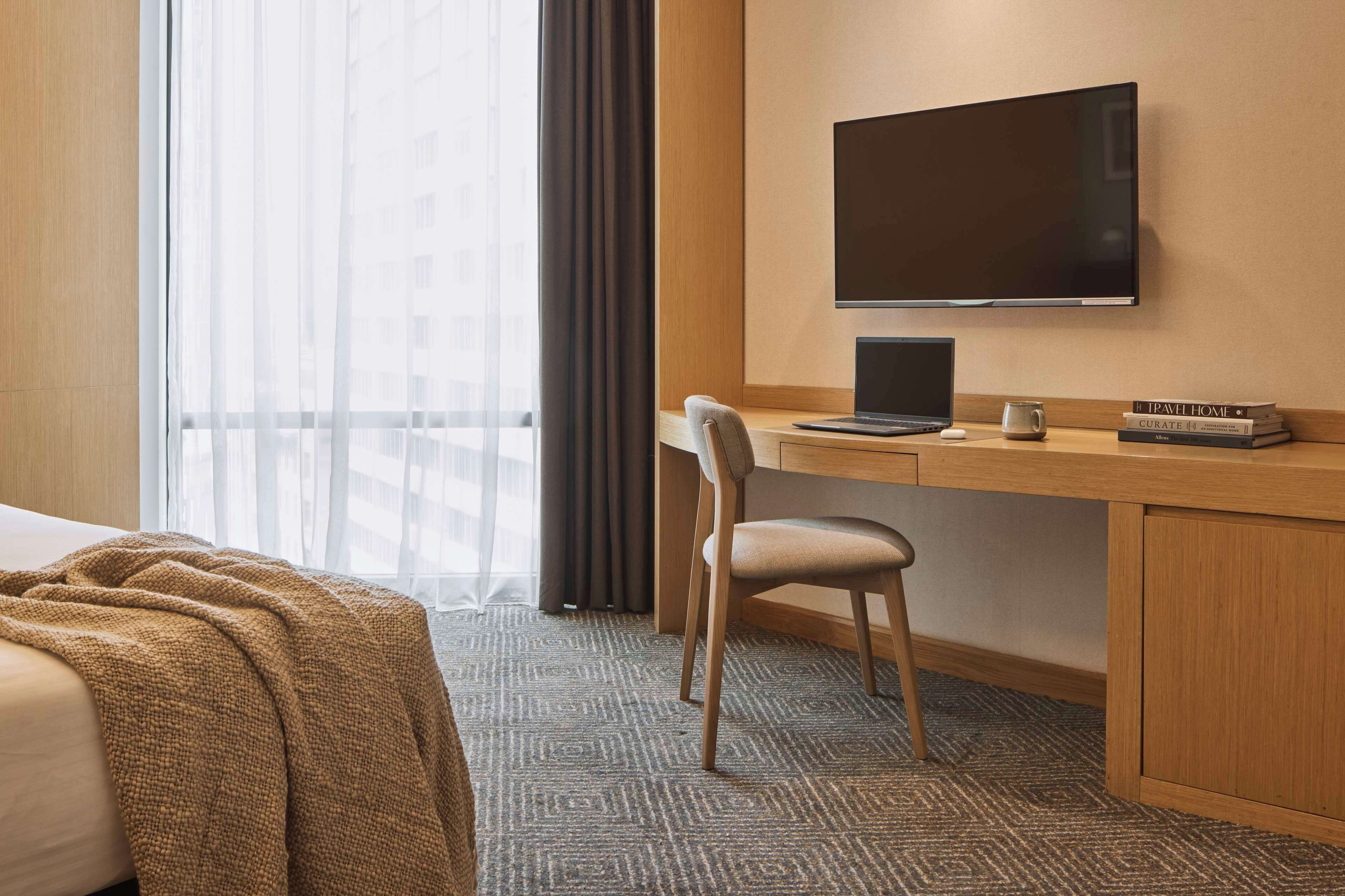 A hotel room with a bed, a desk with a laptop, a TV, and a window with curtains.
