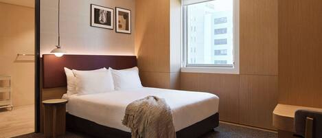 A hotel room with a large bed, a bedside table, a chair, a window with a view of buildings, and framed pictures on the wall.