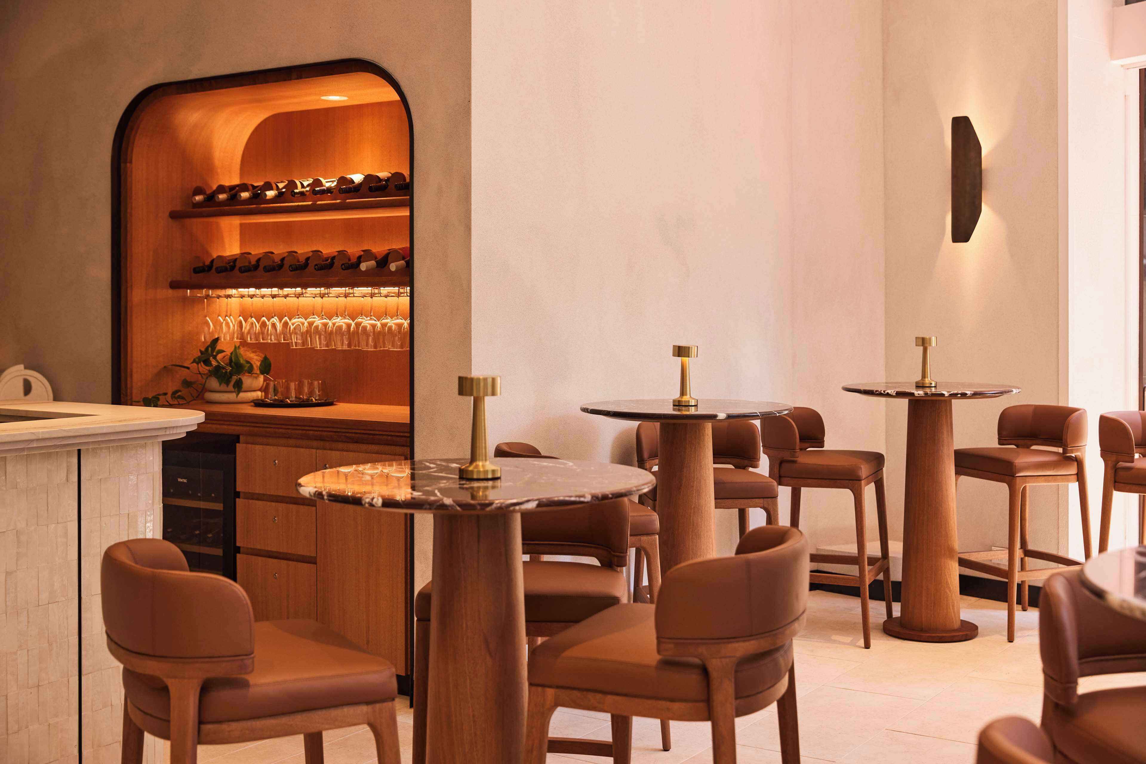 A restaurant interior with round tables and brown chairs, a wine rack, and a bar area.