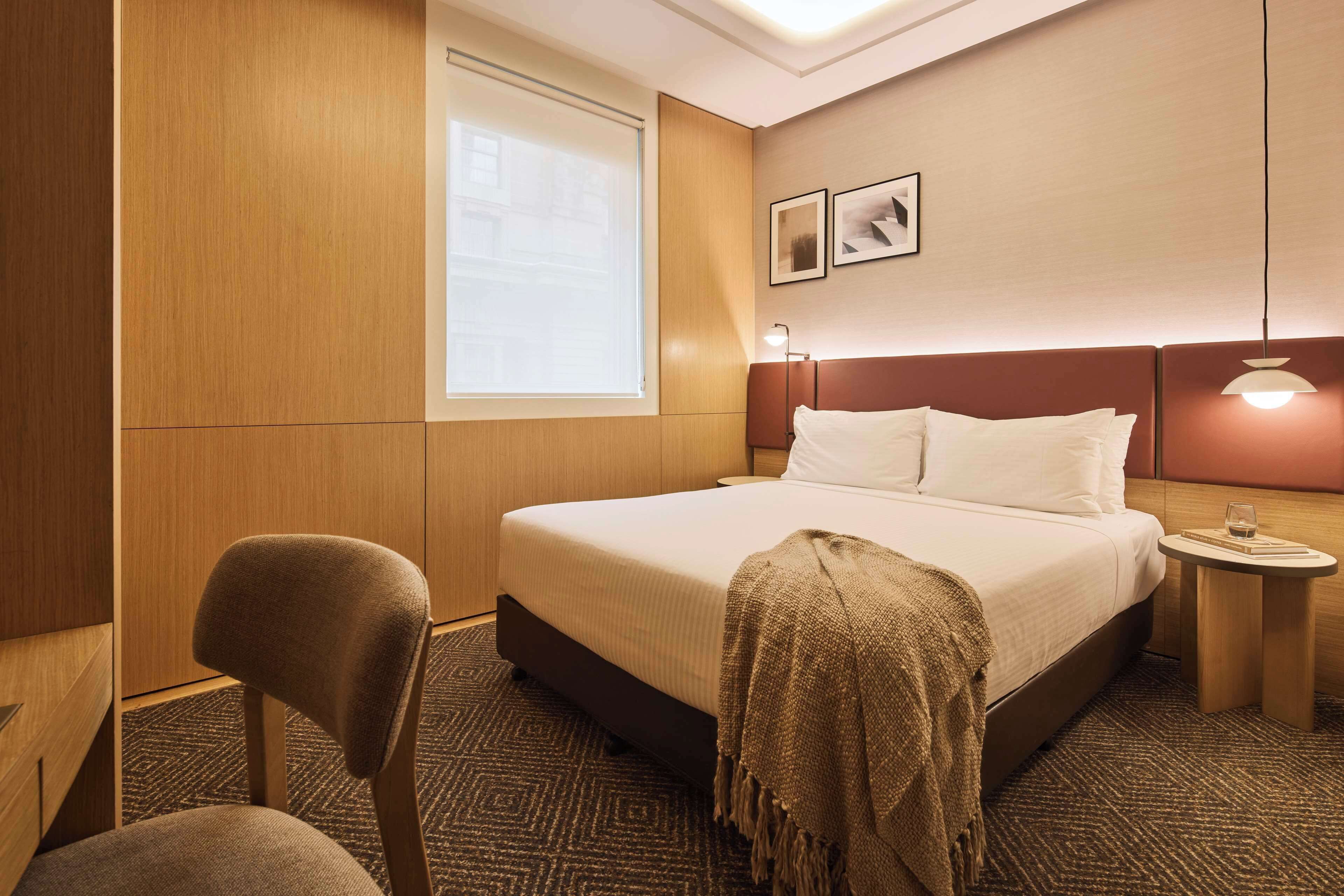 A hotel room with a bed, a chair, a small table with a lamp, and a window with blinds.
