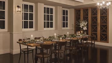 A dining room with a long table set for six, flanked by large windows and a wine cellar visible through a glass door.
