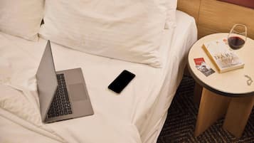 A bed with white linens, a laptop, a smartphone, a glass of red wine, and a book on a small round table beside the bed.