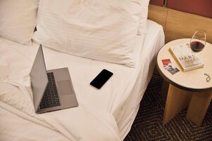 Una cama con sábanas blancas, una laptop, un teléfono inteligente, una copa de vino tinto y un libro sobre una mesita redonda junto a la cama.