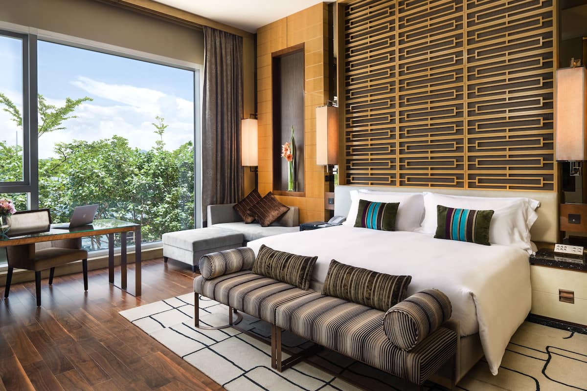 Een moderne hotelkamer met een groot bed, een bank, een bureau met een laptop en uitzicht op groen.