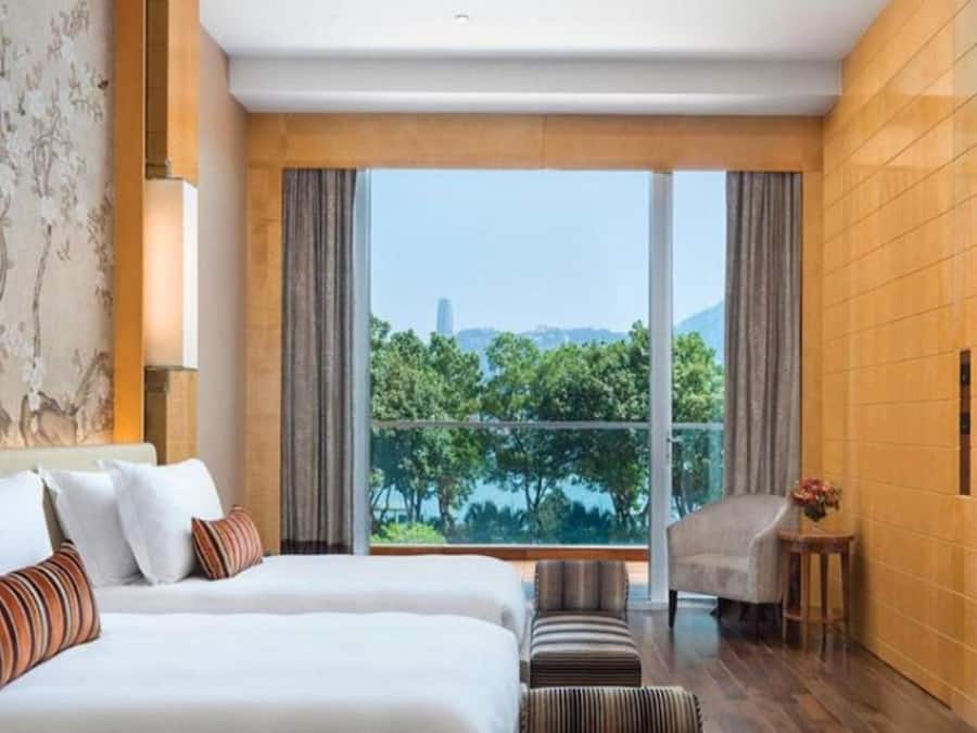 A hotel room with two beds, a view of greenery and a building, and a small seating area.