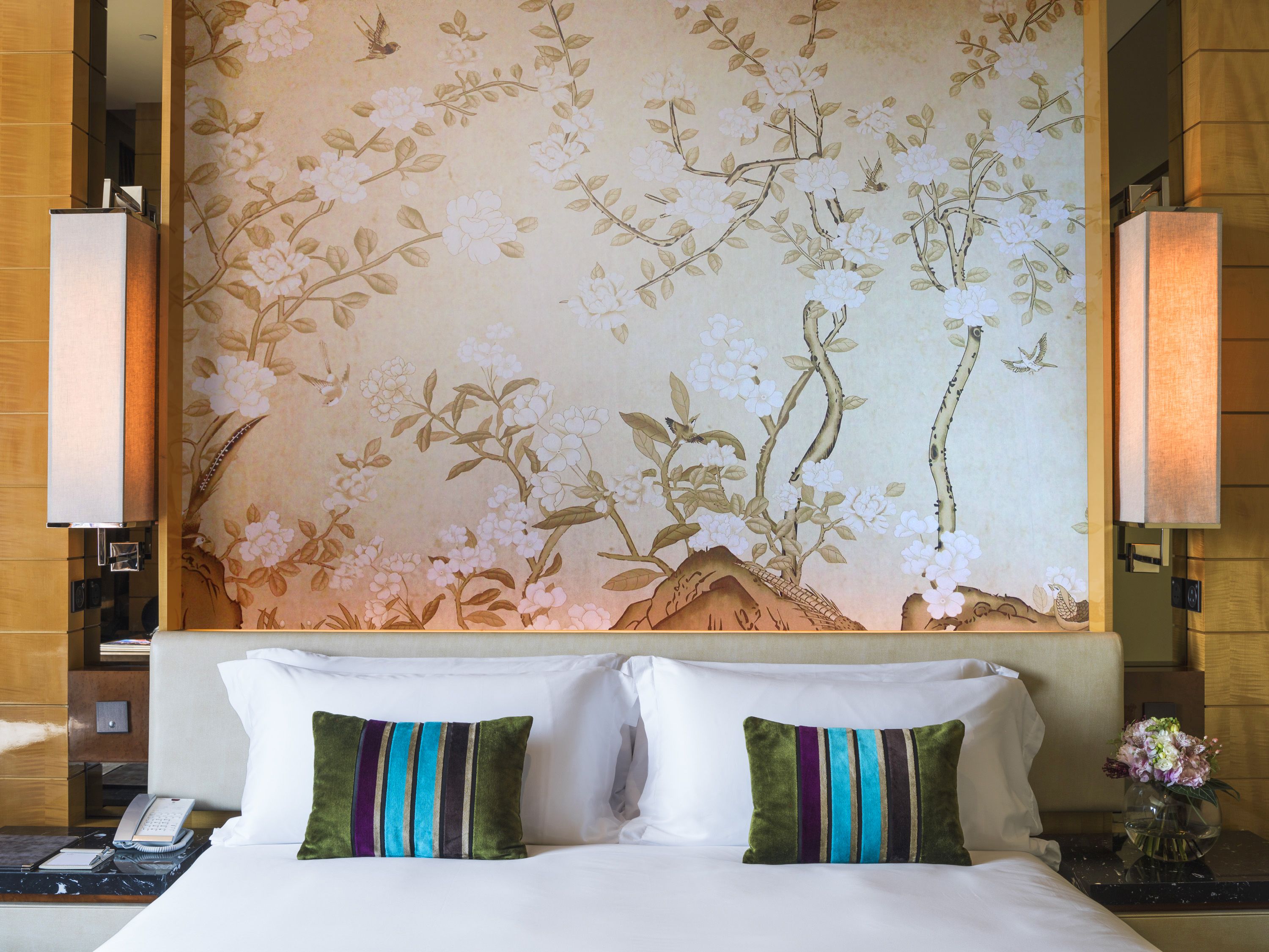 A hotel room with a bed, two pillows, a nightstand, a telephone, and a wall with floral wallpaper.