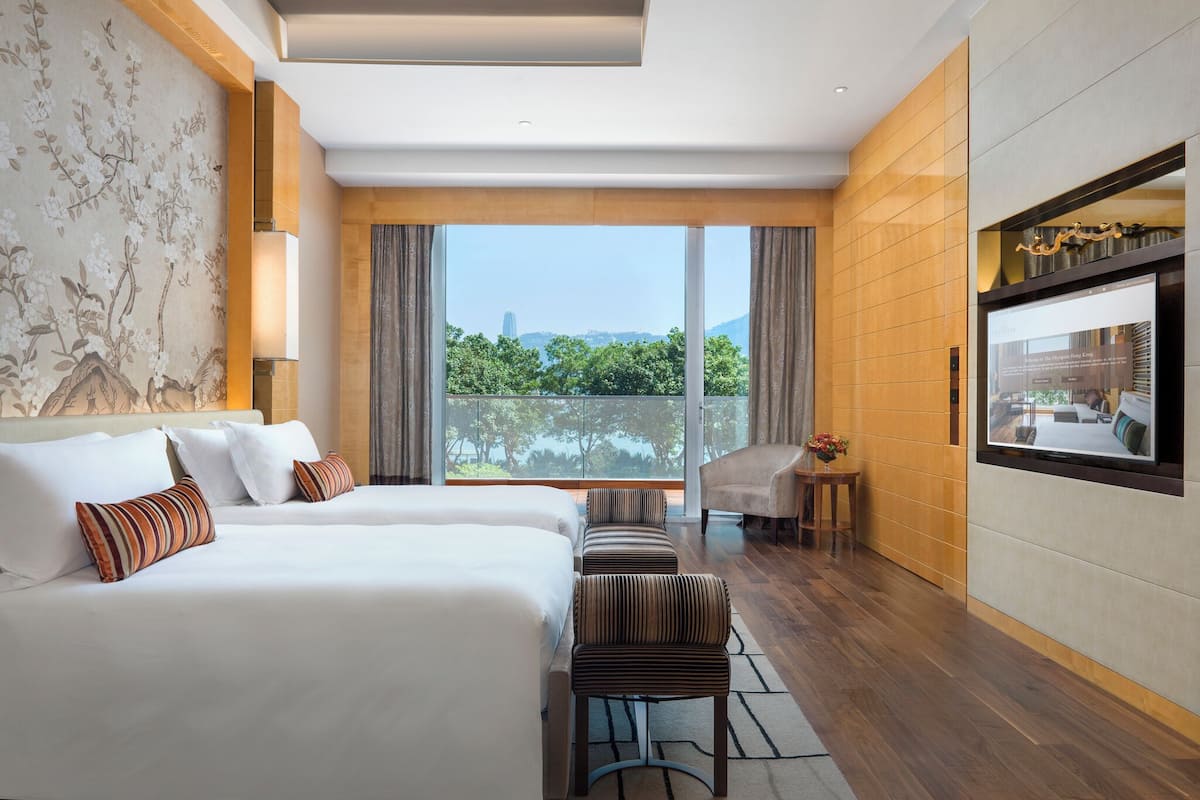 Hotelkamer met een groot bed, een kleiner bed, een bank, een stoel, een televisie en een balkon met uitzicht op groen en een gebouw.