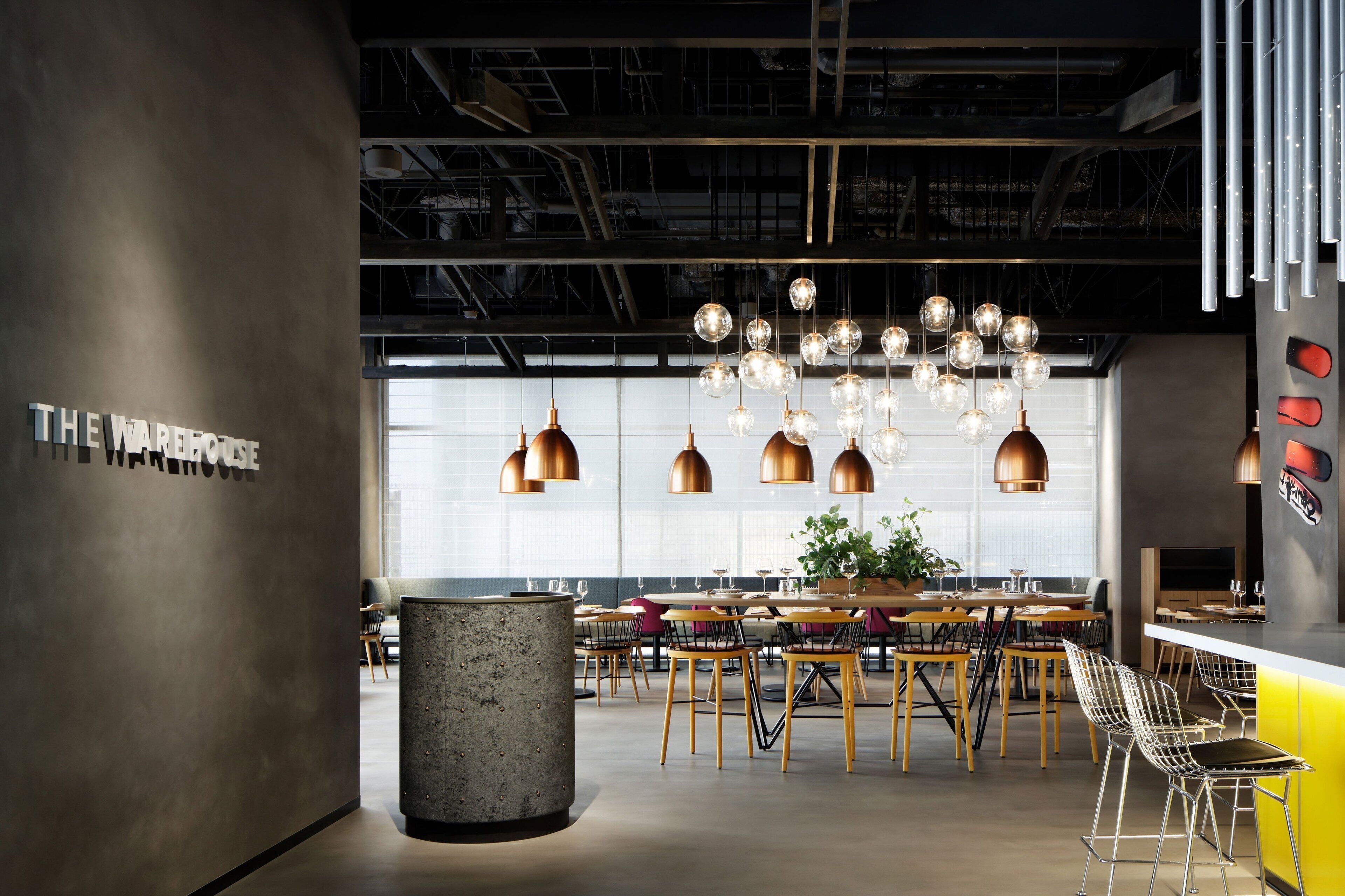 A modern restaurant interior with a central dining area, hanging spherical light fixtures, and a sign reading "THE WAREHOUSE".