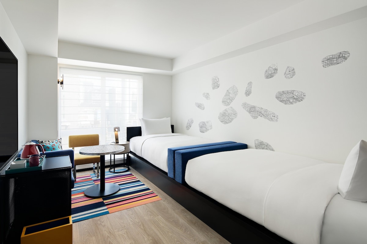 A modern hotel room with a large bed, a small desk, a chair, and a window with blinds.