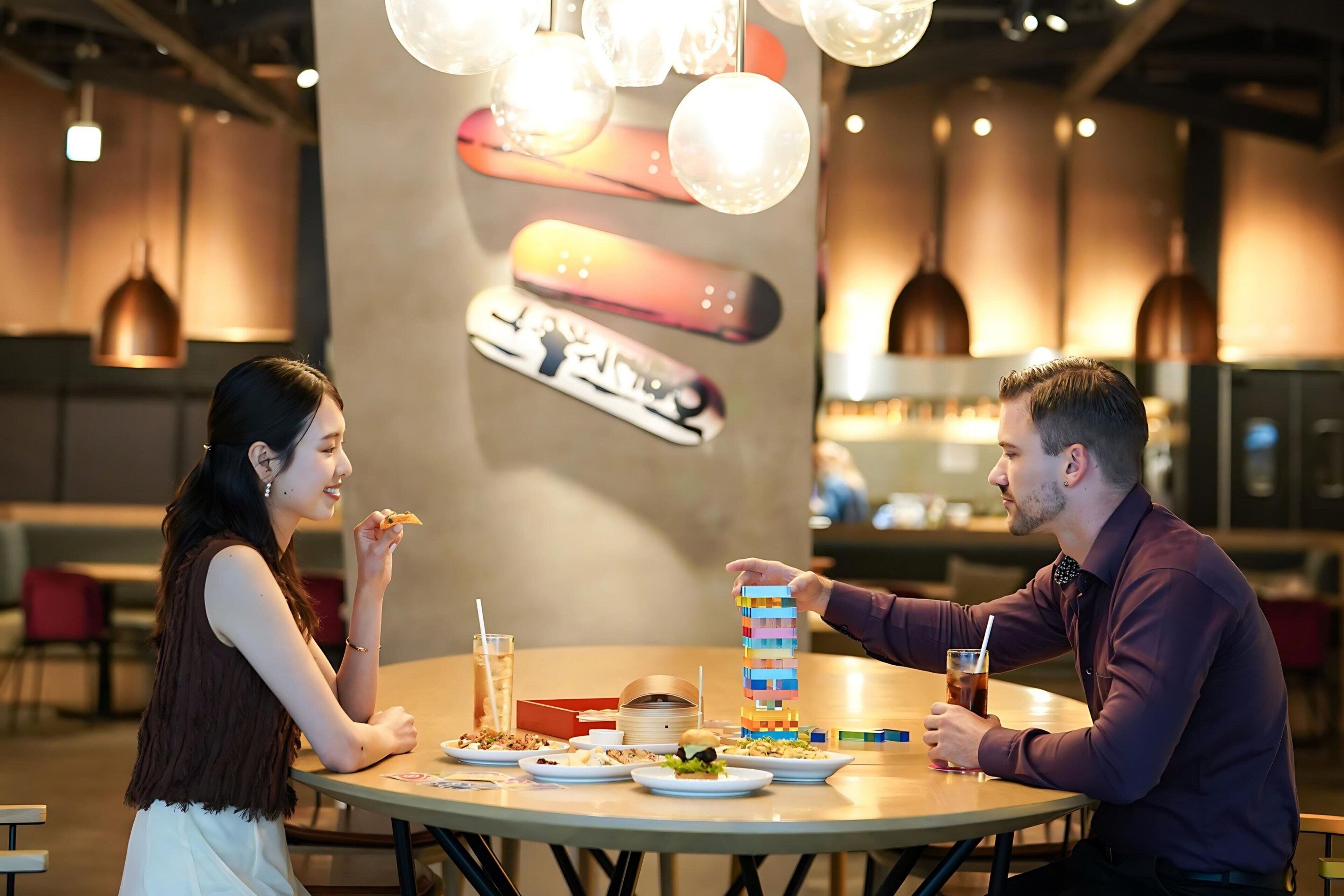 A restaurant setting with a table set for two, featuring plates of food, a drink, and a jenga-like game.