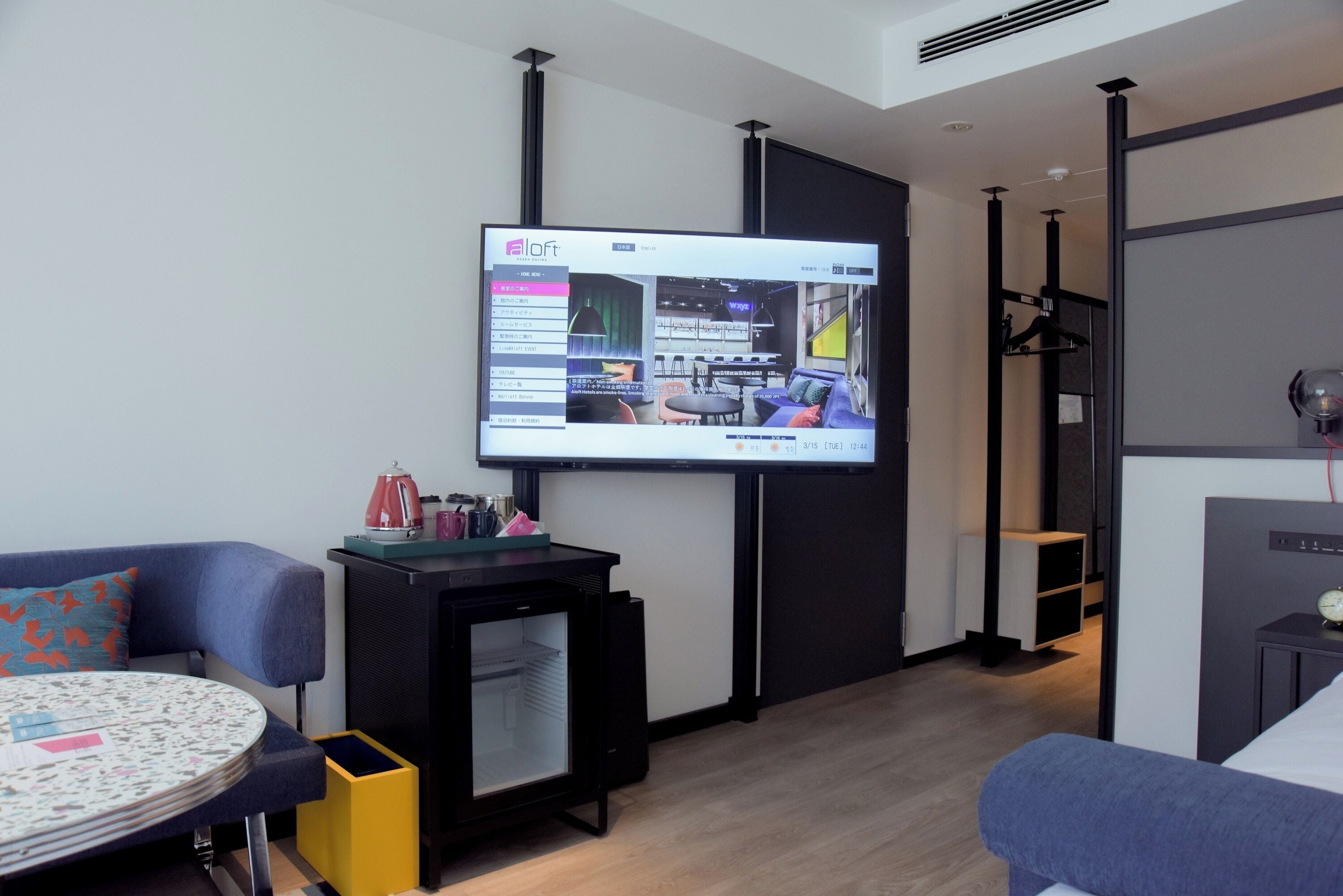 A modern hotel room with a large flat-screen TV mounted on the wall, a small black refrigerator, a round table with a floral tablecloth, and a blue sofa.