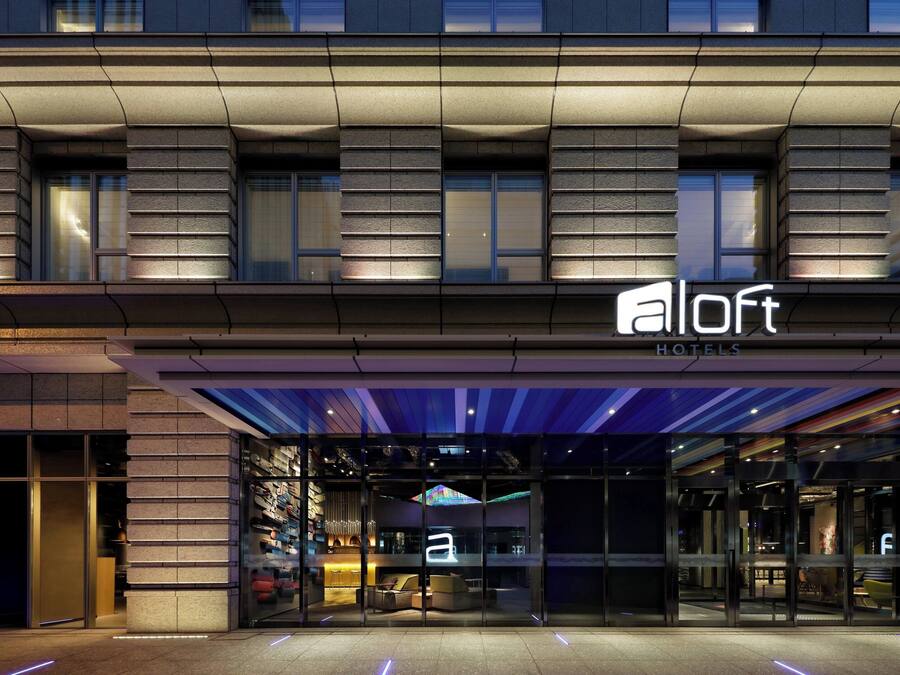 Una entrada de hotel moderna con fachada de vidrio, un mostrador de recepción y un área de estar interior visible.