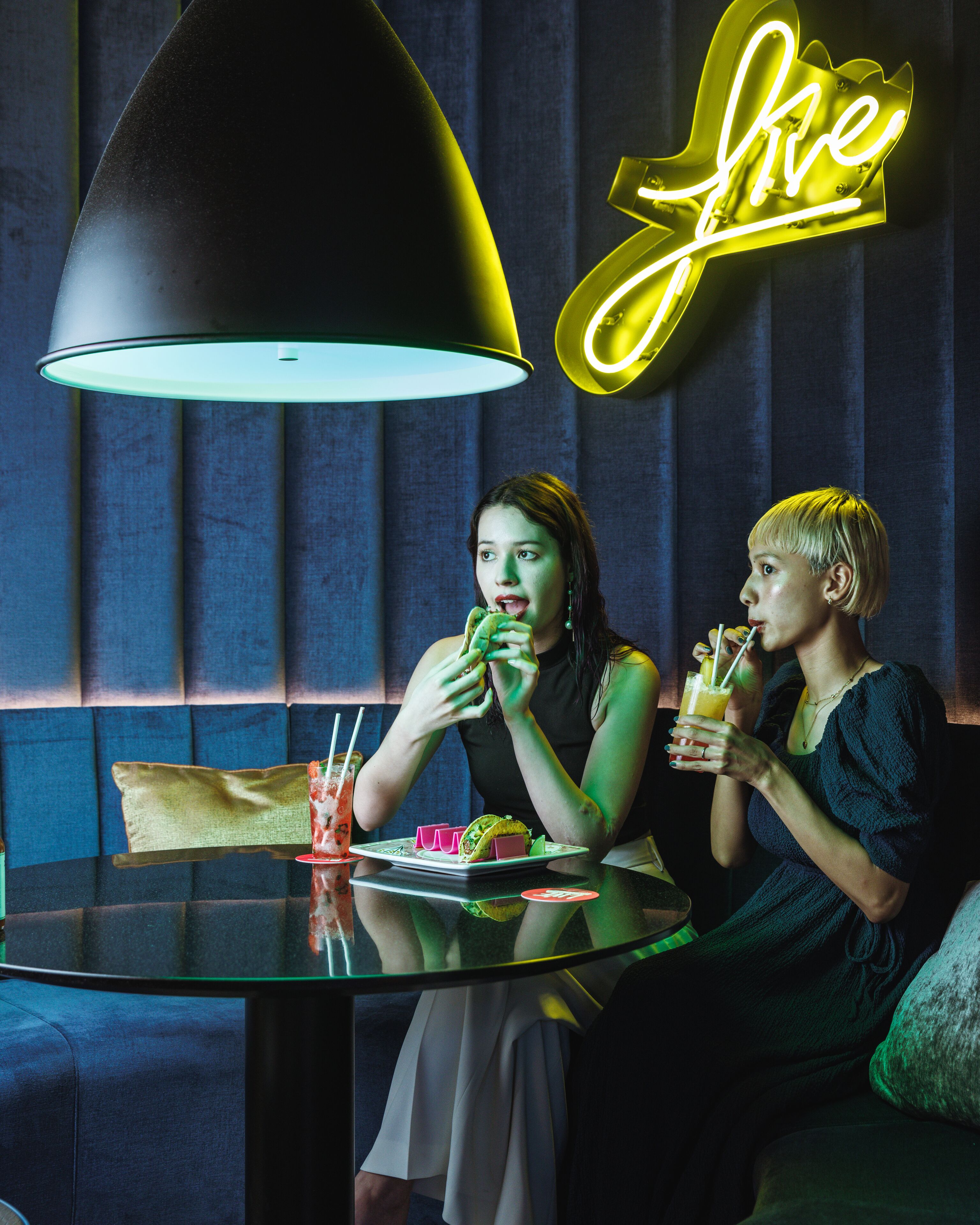 Two people dining at a table with a neon sign in the background.