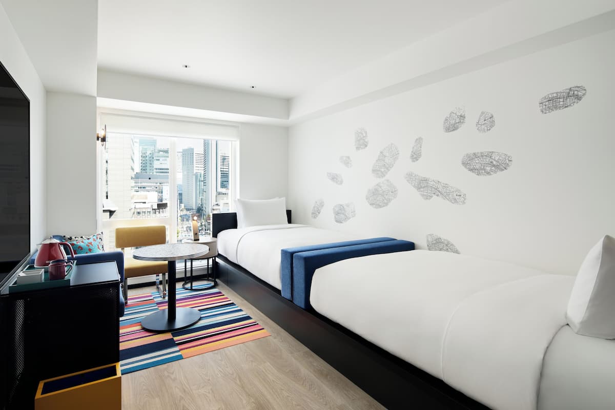 A hotel room with a large window, a bed, a desk, a chair, and a colorful striped rug.