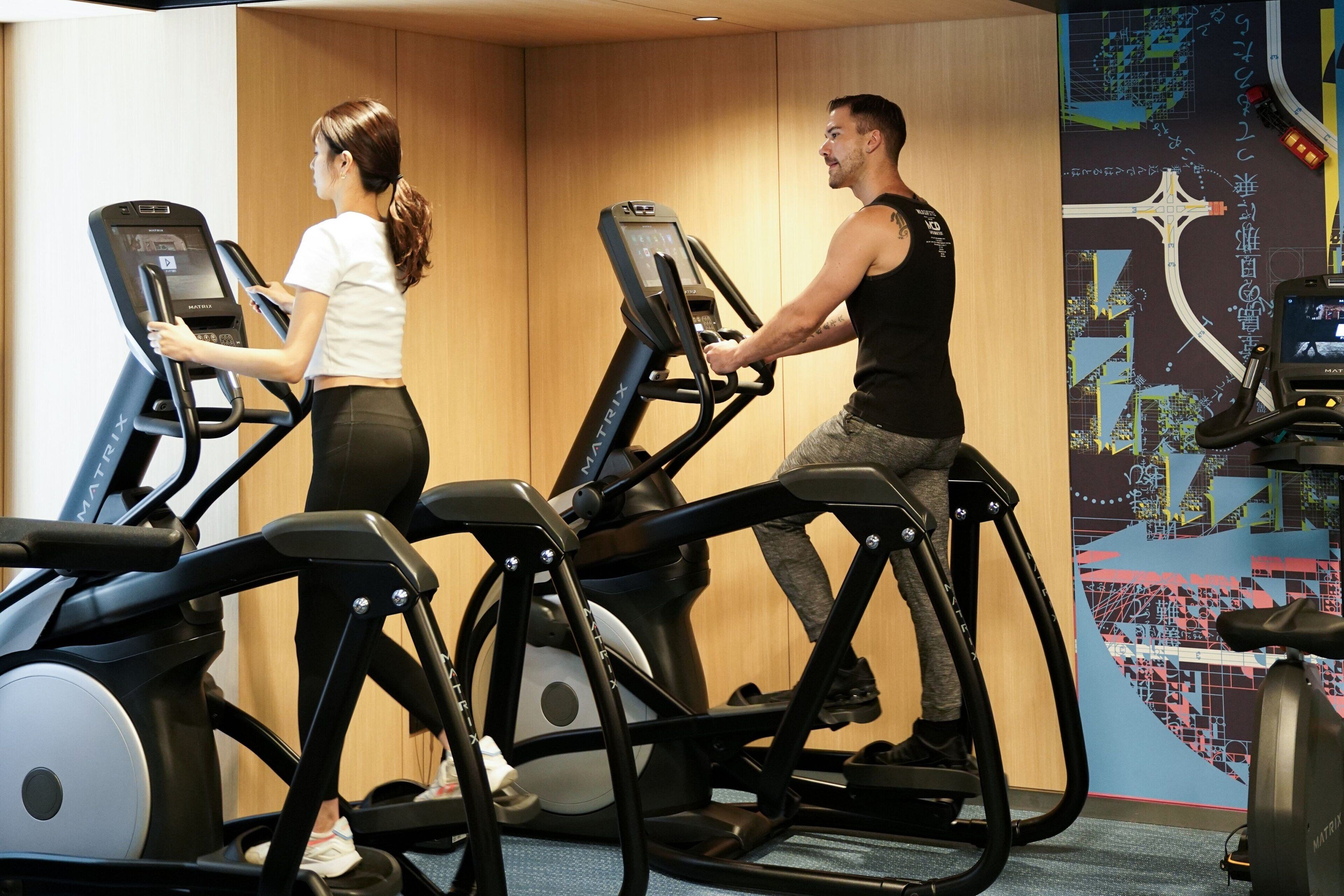 Två personer som använder löpband i ett gym, med en stor väggmålning i form av en karta.