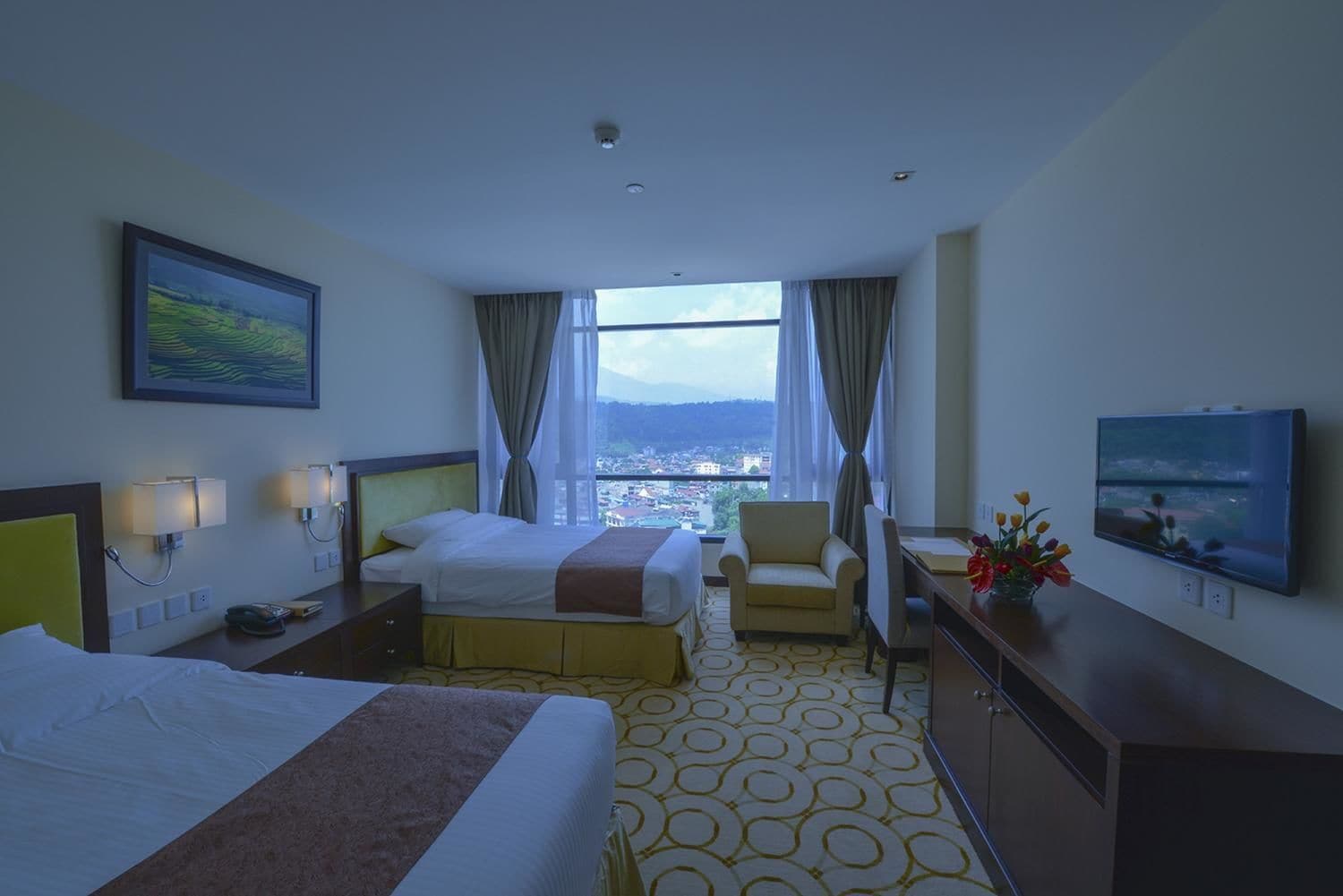 Habitación de hotel con dos camas, un escritorio, una silla, un televisor y un ventanal con vistas a la ciudad.