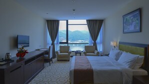 Una habitación de hotel con una cama, dos sillones, un escritorio con televisor y vistas a un cuerpo de agua y montañas.