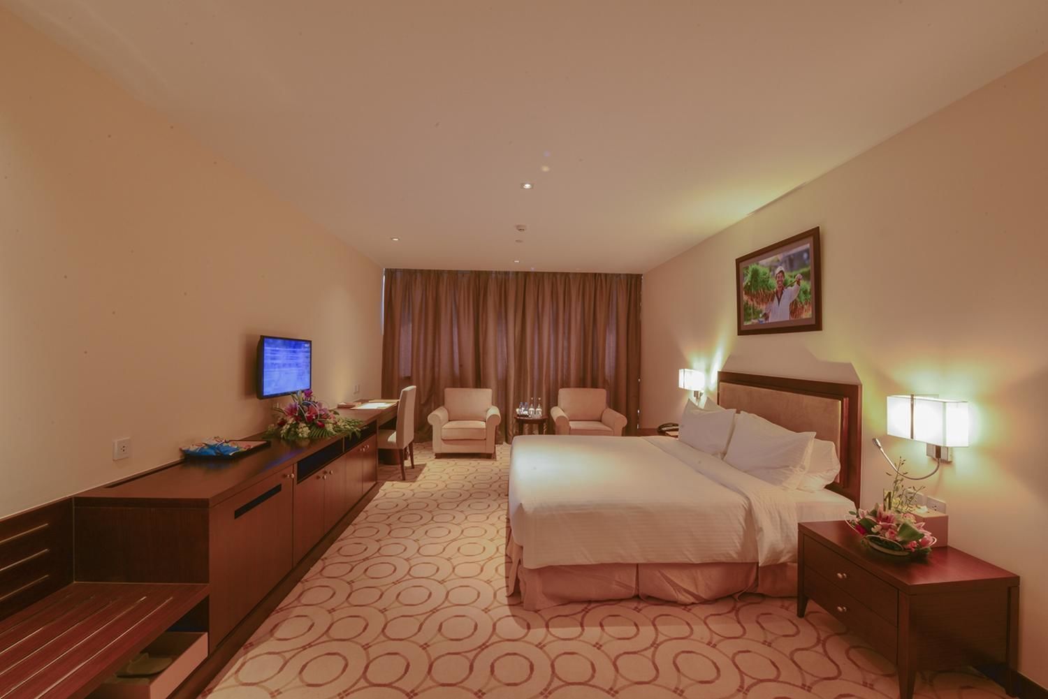 Een hotelkamer met een bed, een bureau met een computer, twee fauteuils, een televisie en een schilderij aan de muur.