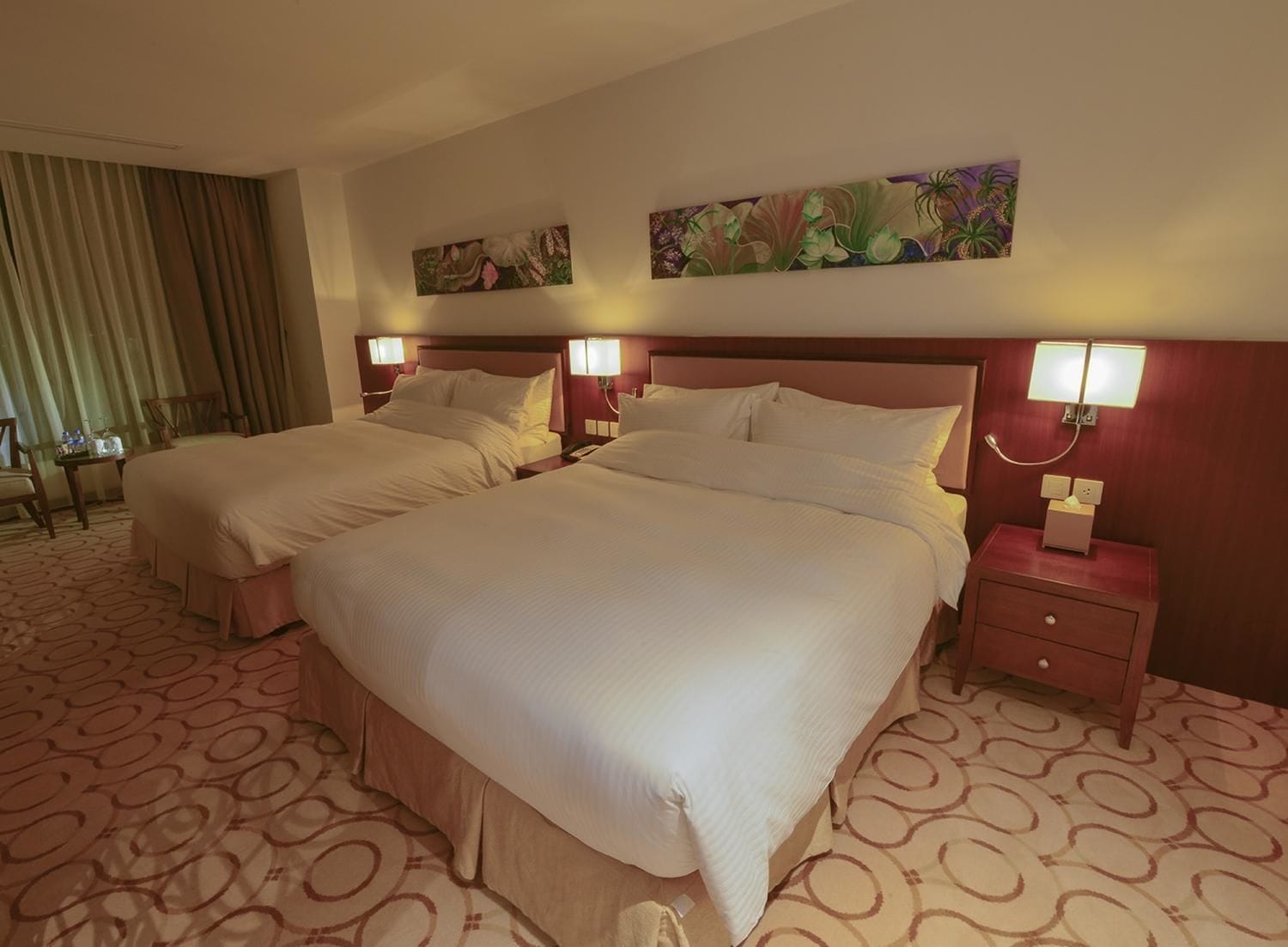 A hotel room with two beds, a desk, and a chair. There are paintings on the walls and a patterned carpet.
