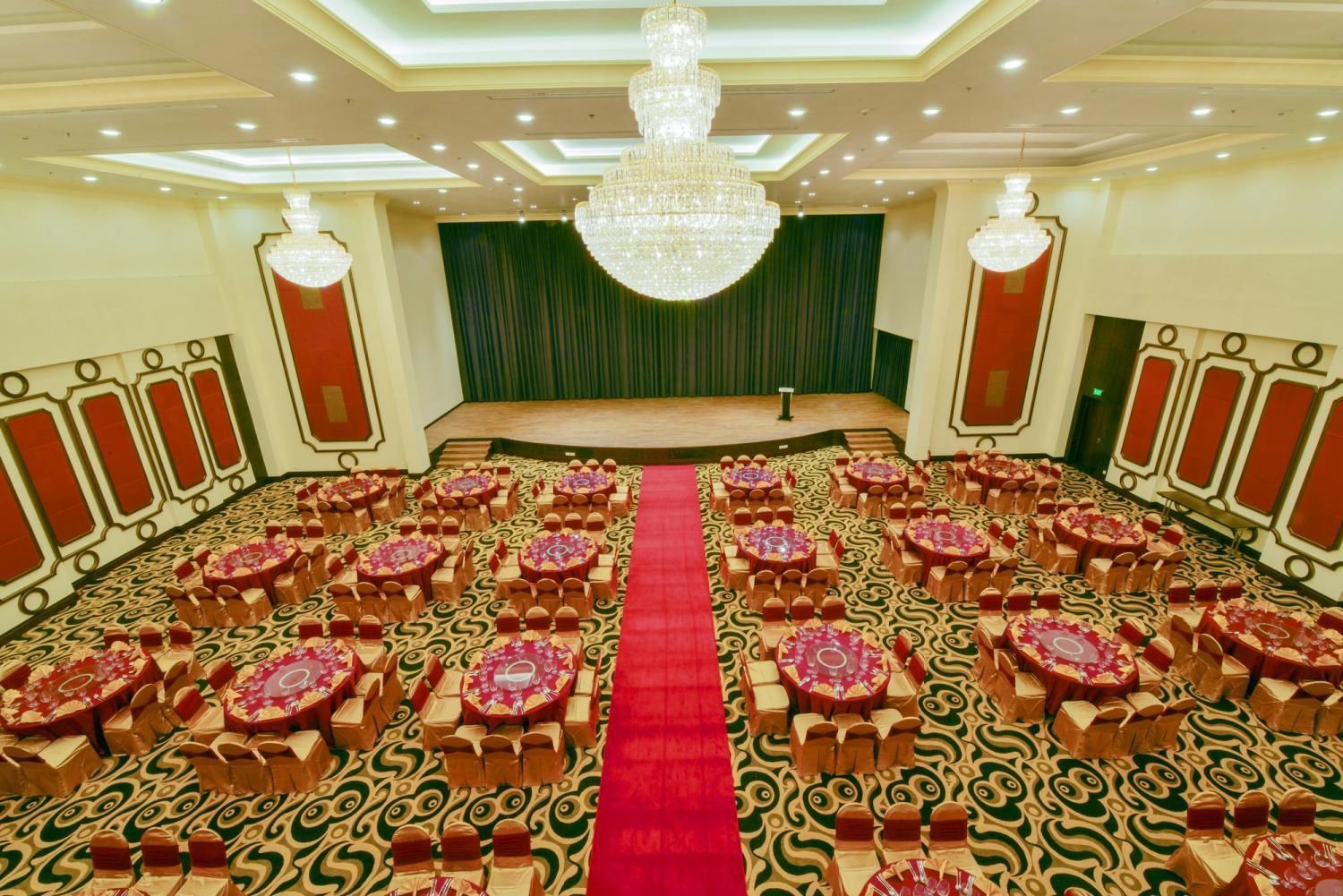 Un salón de conferencias con un pasillo central, filas de mesas con sillas, una alfombra roja y una araña.