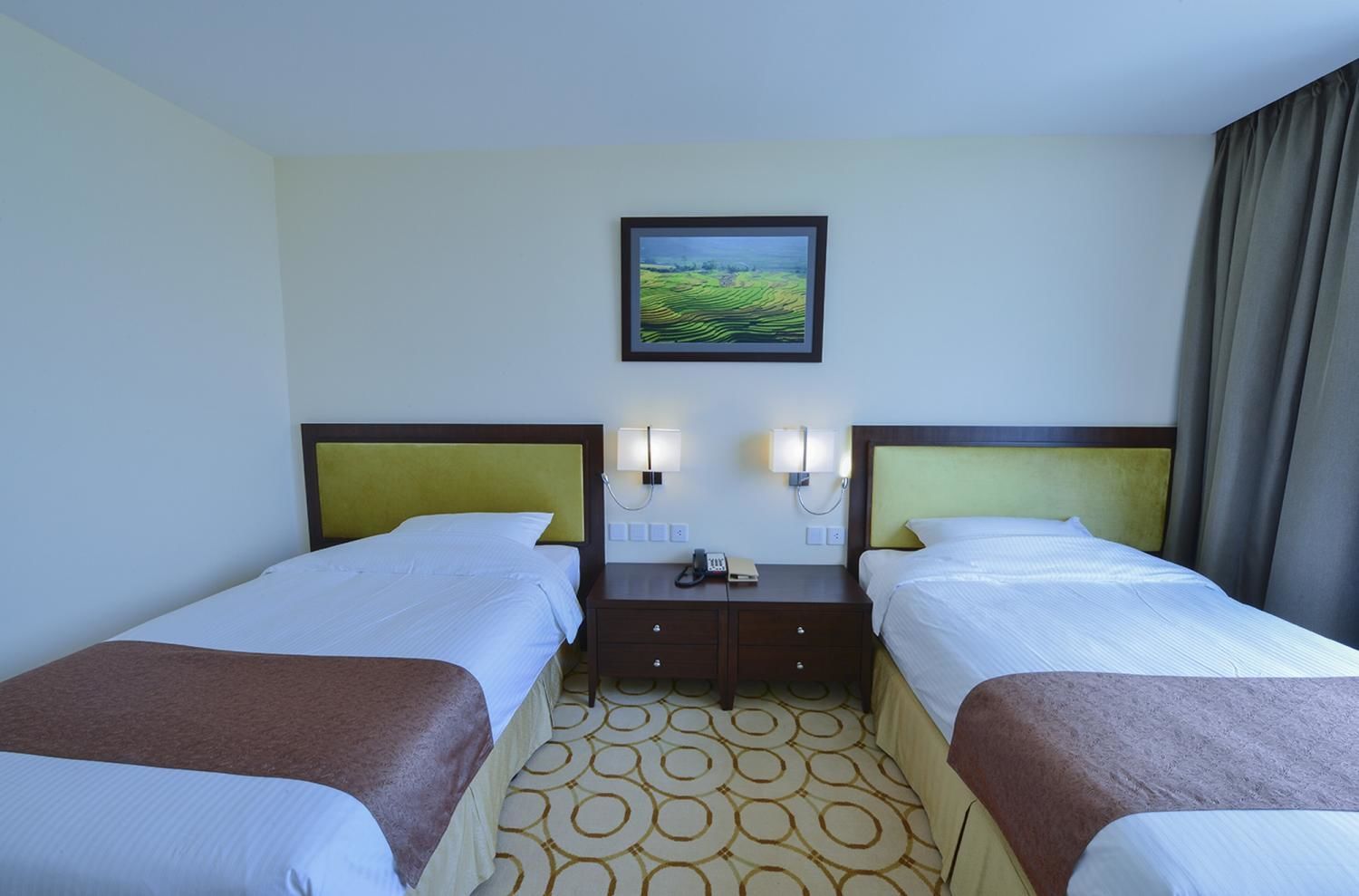 A hotel room with two beds, a nightstand with a lamp, a framed picture on the wall, and a patterned carpet.