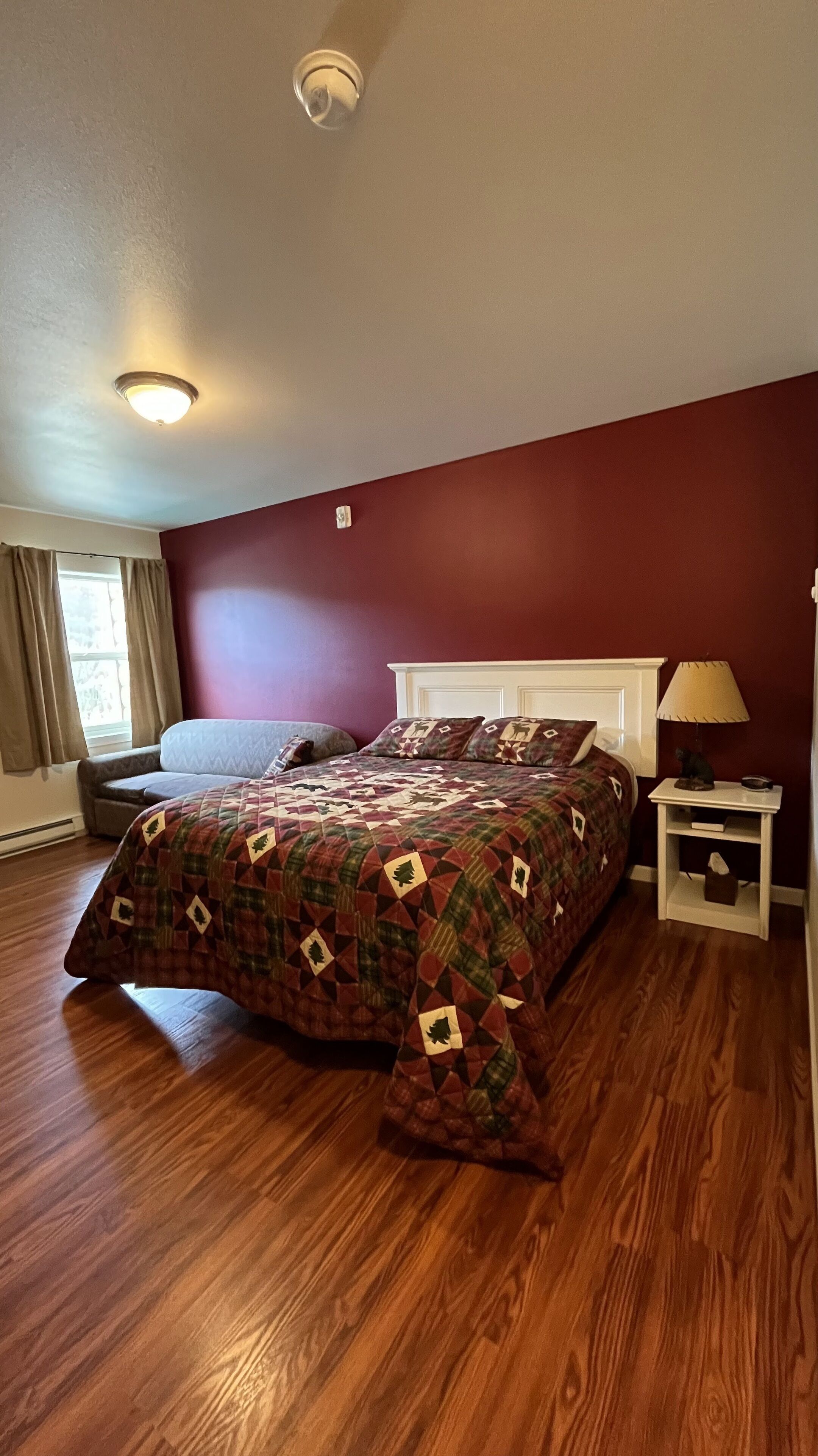 Une chambre avec un plancher en bois, un lit recouvert d’une courtepointe à motifs, une table de chevet avec une lampe et une fenêtre avec des rideaux.