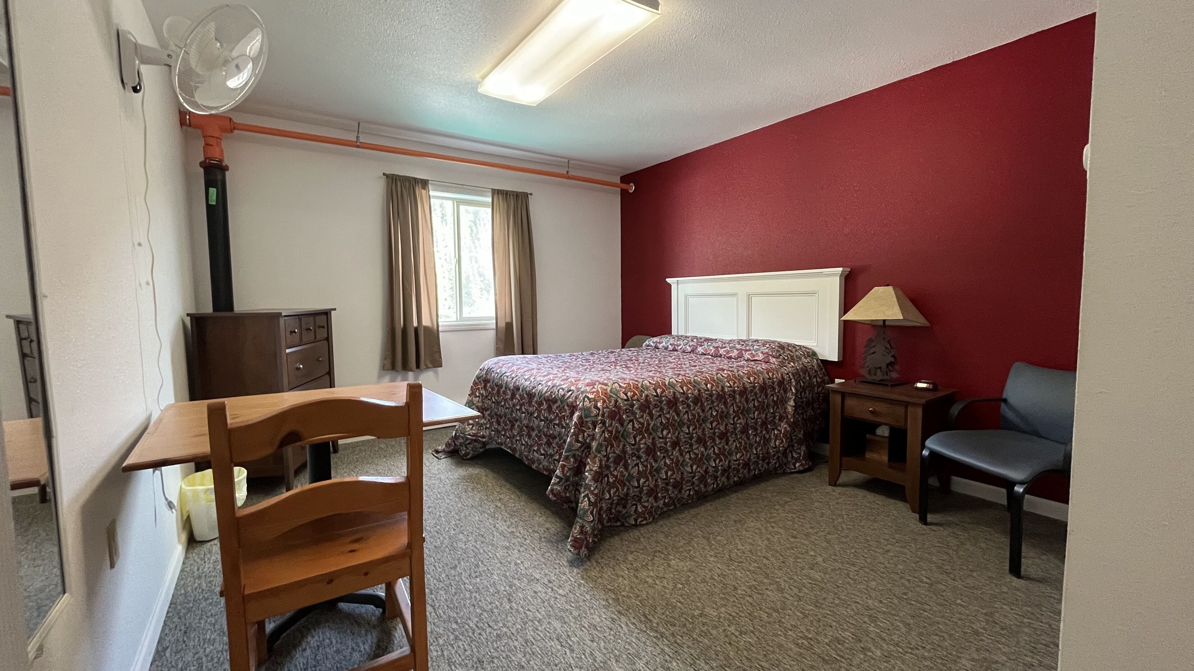 A hotel room with a bed, a desk, a chair, a small table with a lamp, a fan, and a window with curtains.
