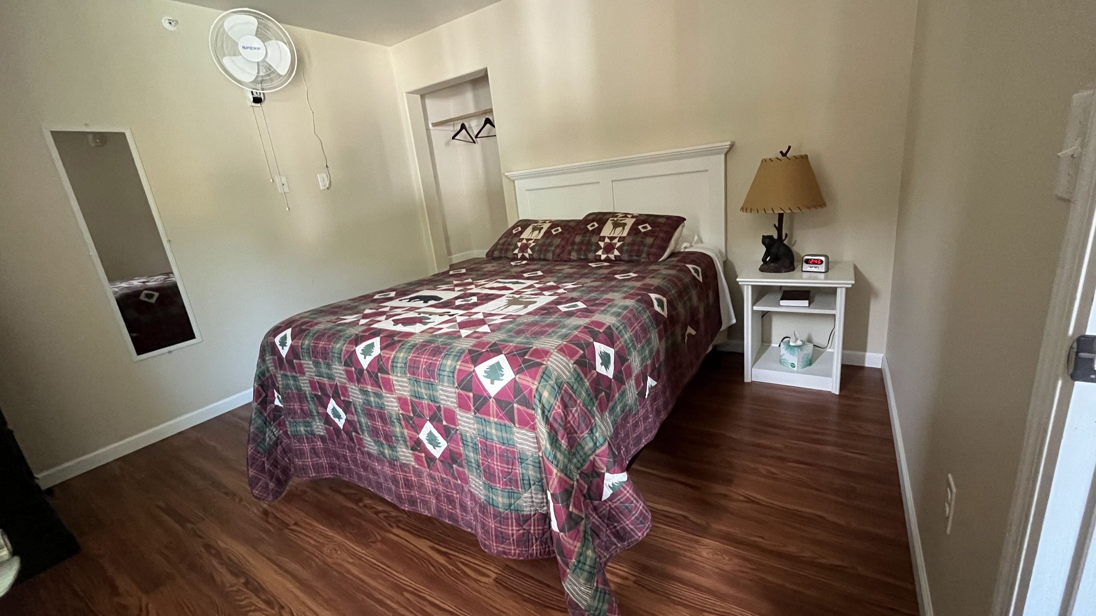 Un dormitorio con cama, mesita de noche, lámpara, reloj y espejo en la pared.