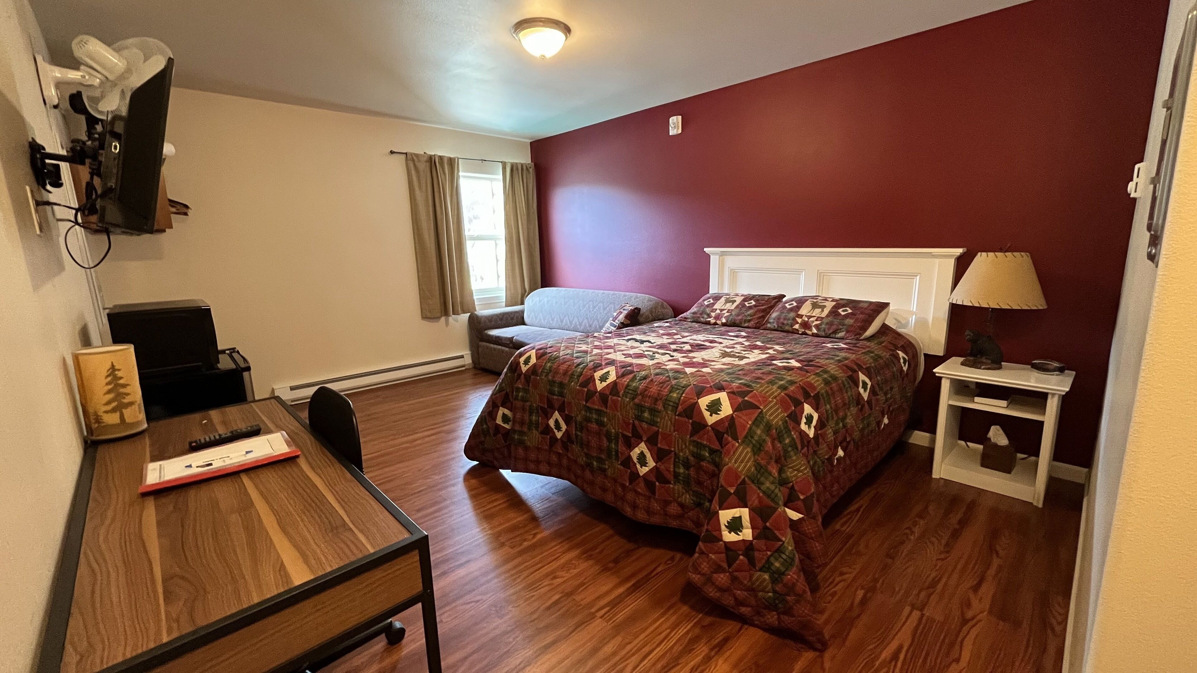 Un dormitorio con cama, escritorio, silla, mesita de noche, lámpara y ventana con cortinas.