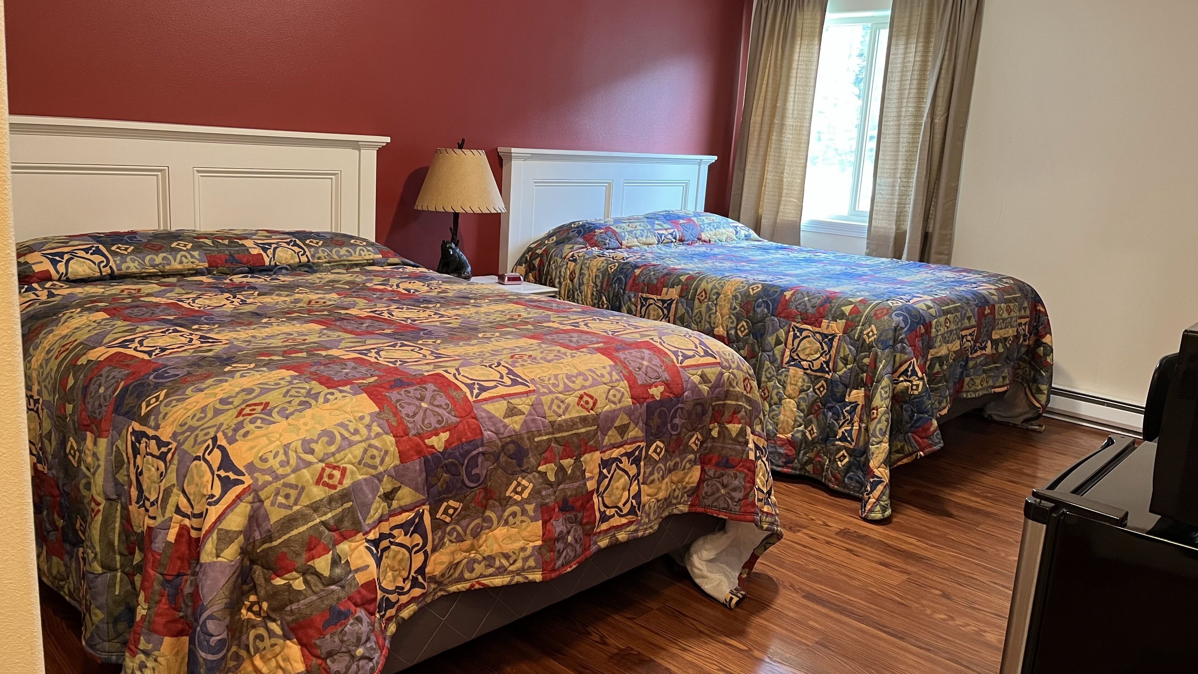 A room with two beds, each covered with a colorful quilt. There is a lamp on a nightstand, a window with curtains, and a TV on a stand.