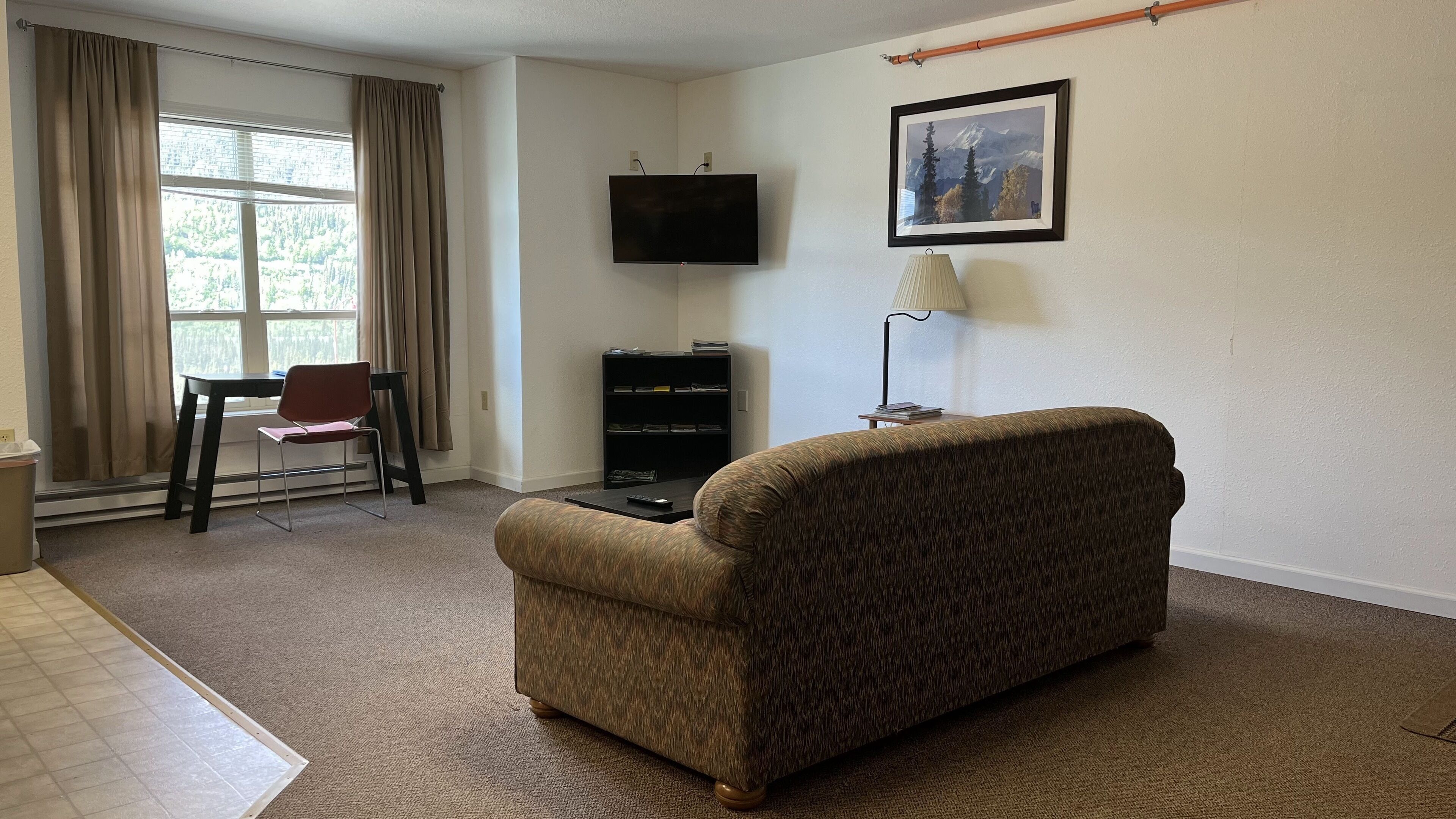 A living room with a sofa, a TV mounted on the wall, a bookshelf, a chair, and a window with curtains.