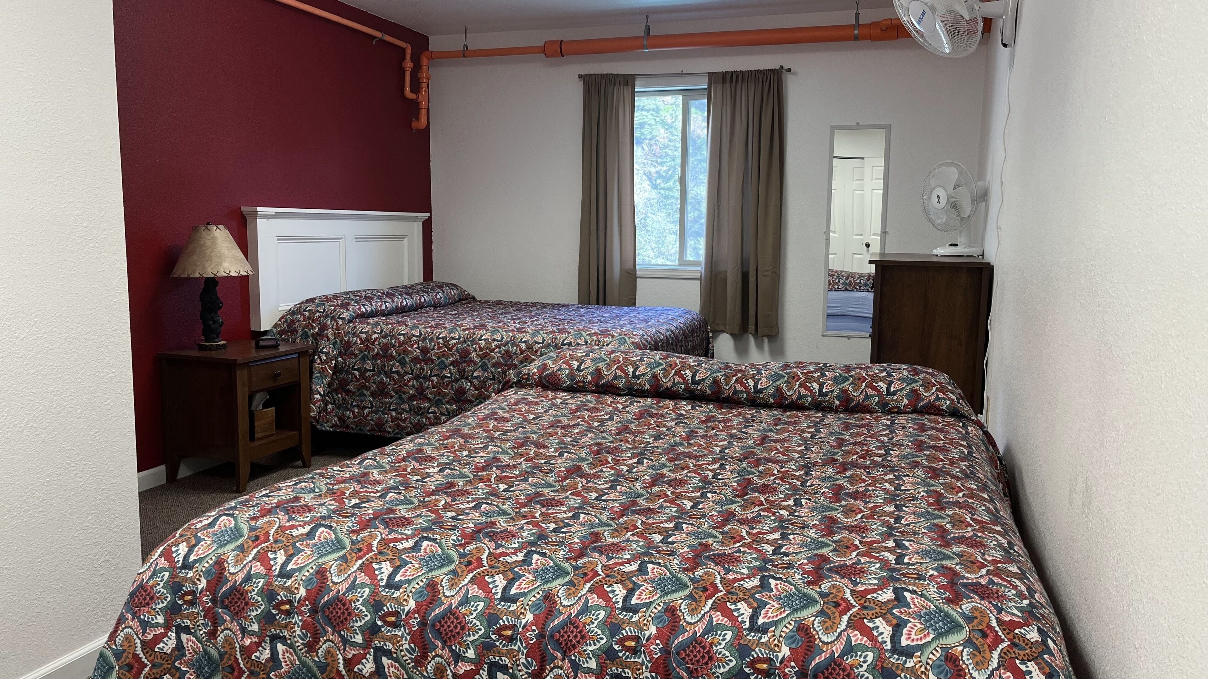 A room with two beds, a nightstand, a lamp, a window with curtains, and a fan.
